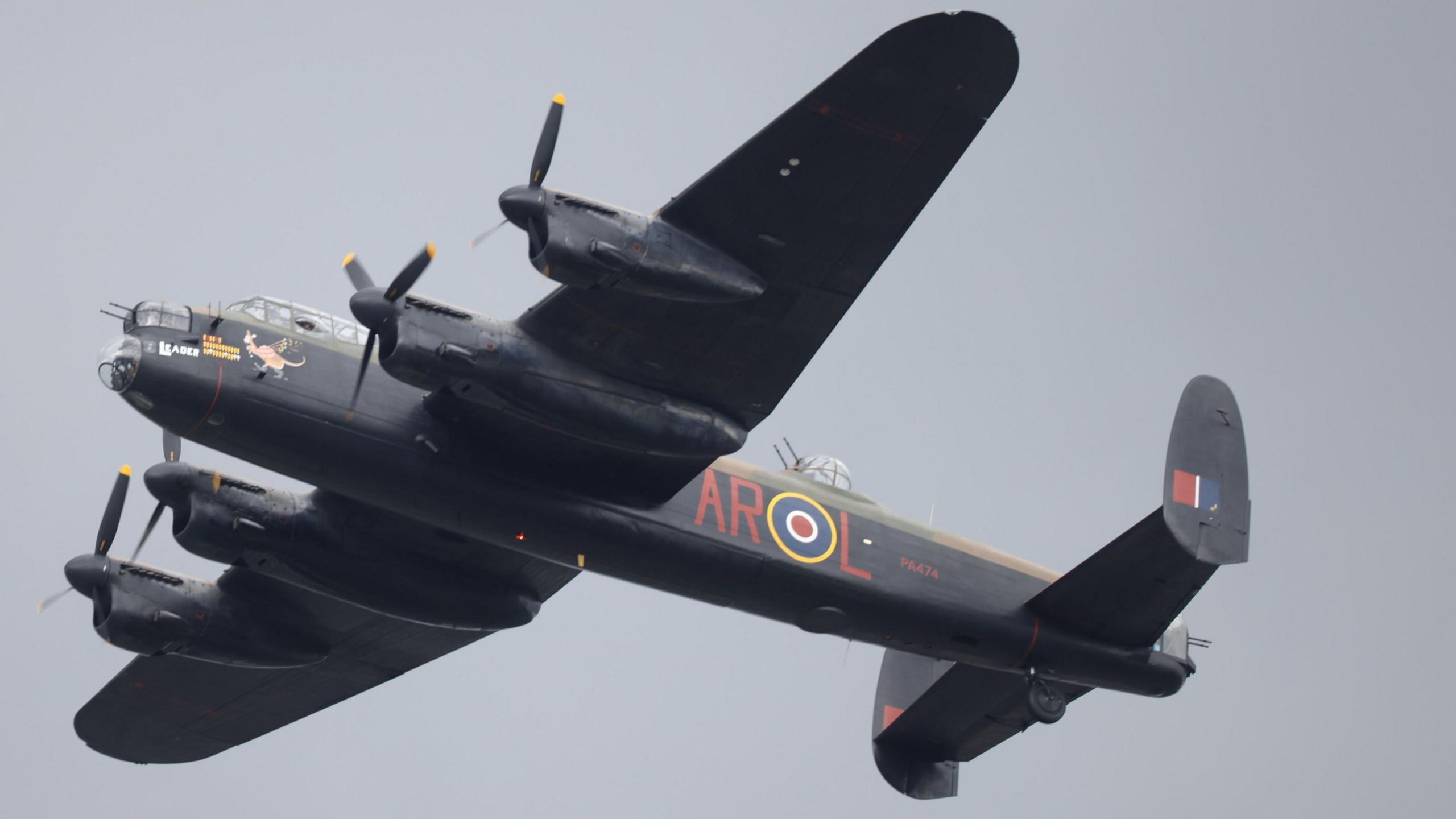 A Lancaster bomber aircraft flying in a cloudy sky. Its wings have two propellers each and the aircraft is black with the word AROL in red lettering on the side.