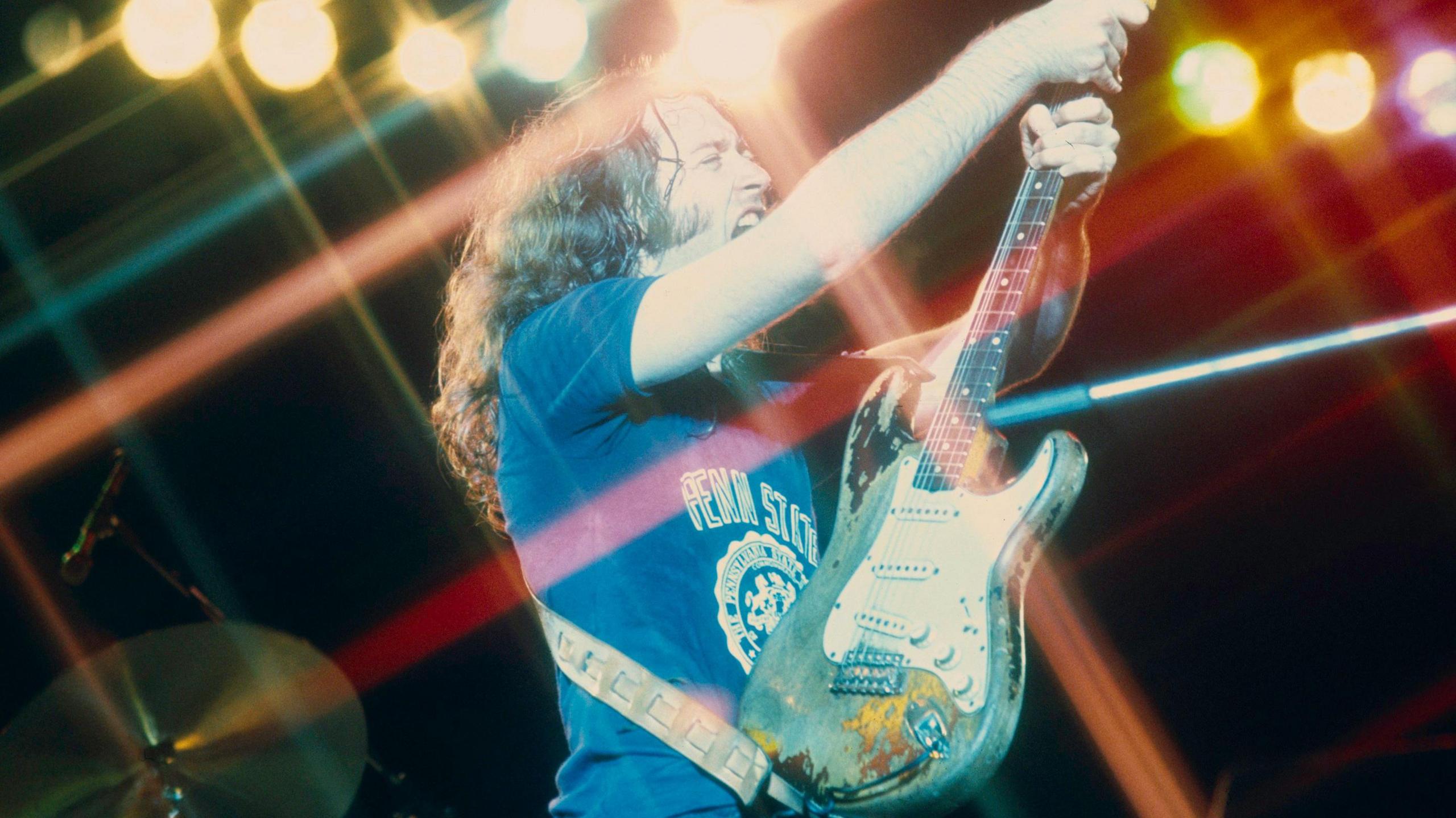 Musician Rory Gallagher holds aloft a fender stratocaster guitar, he is wearing a blue t-shirt, and drums can be seen behind him. He has long hair and is standing is in a number of spotlights