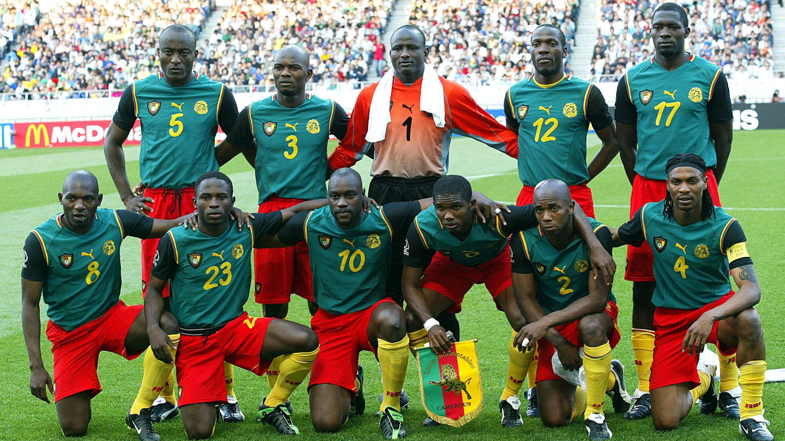 A Cameroon team photograph taken on the pitch before a game at the 2002 World Cup. Six players are down on one knee or on their haunches in the front row, with captain Samuel Eto'o, third from the right, holding a team pennant. The goalkeeper is in the centre of five players standing in the back row and has a white towel draped around his neck. The outfield players wear a kit made up of yellow socks, red shorts and a green sleeveless shirt which has had black upper-arm sleeves sewn on to it. The goalkeeper sports an orange top and black shorts 