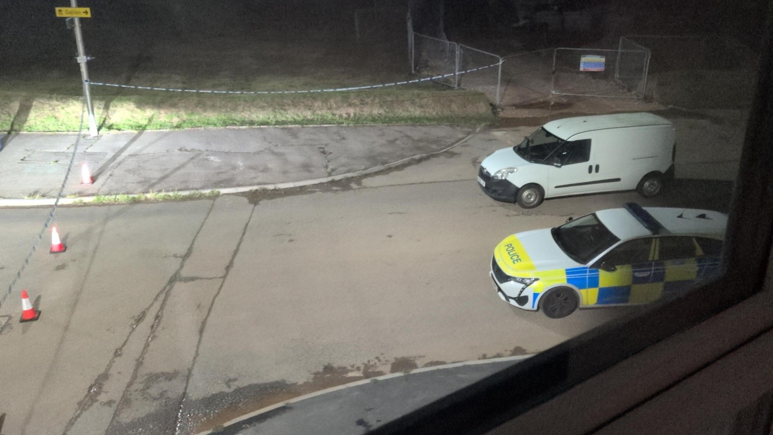 A police car and a white van parked up on a road at the edge of a police cordon following a suspected stabbing. Police tape has been attached to lampposts along the edge of the cordon.