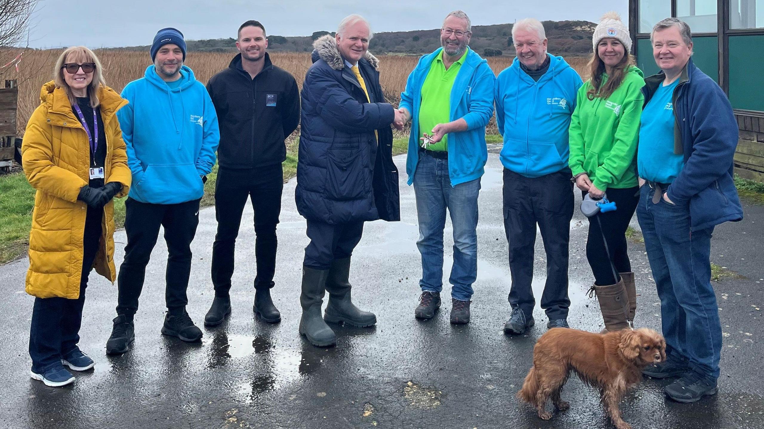 Members of Hengistbury Head Outdoors with Councillor Mike Cox and the BCP Council team posing in a line outside the centre. There are two women and six men in the group, and a brown spaniel. It's a grey, wet day and five of the people are wearing matching brightly-coloured blue or green hoodies.