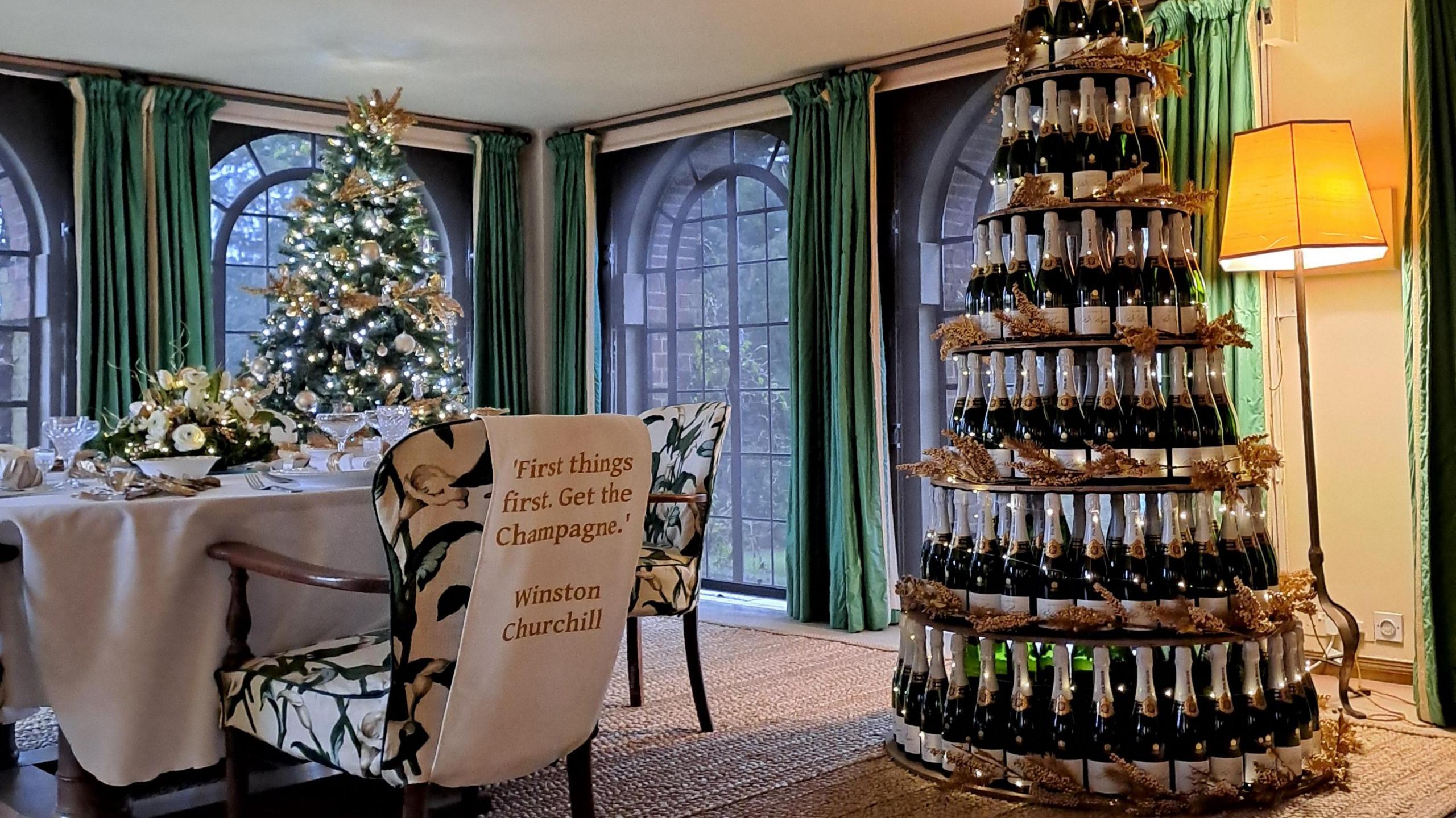 A Christmas tree made up of champagne bottles next to a table with a decoration on a chair saying "First things first. Get the champagne. Winston Churchill."