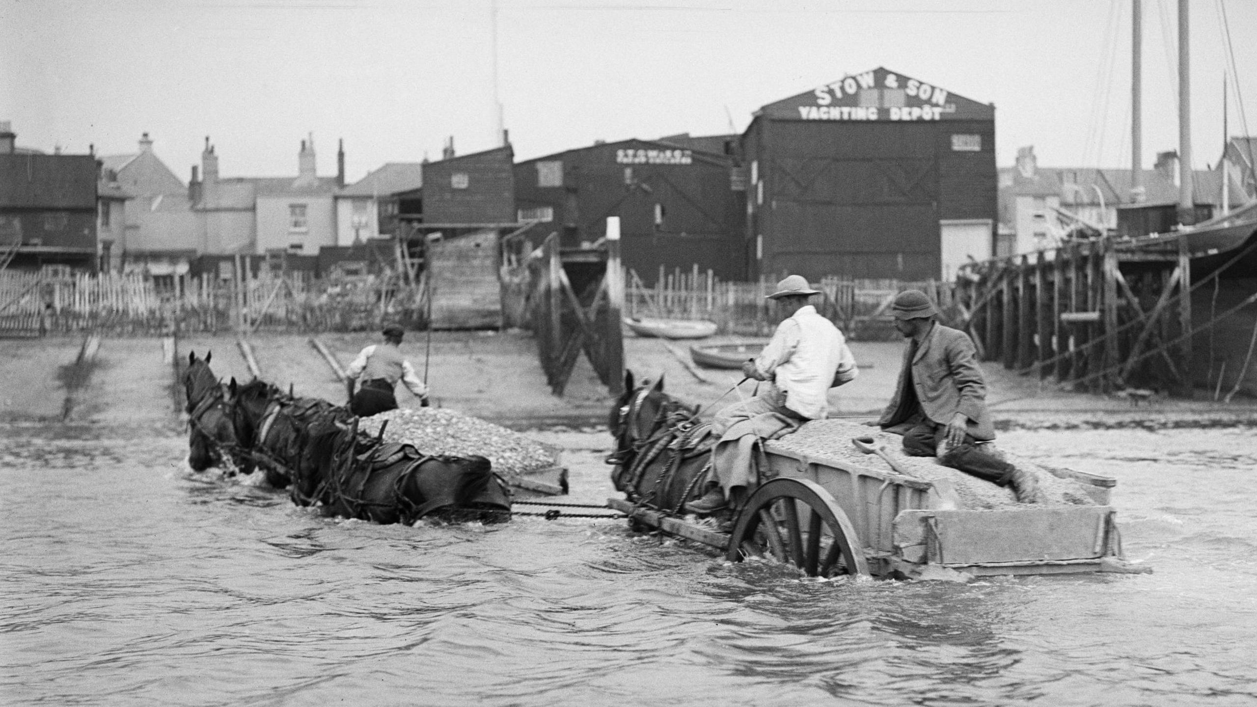 A black and white image of two men in a horse-drawn carriage in the water heading towards a beach.