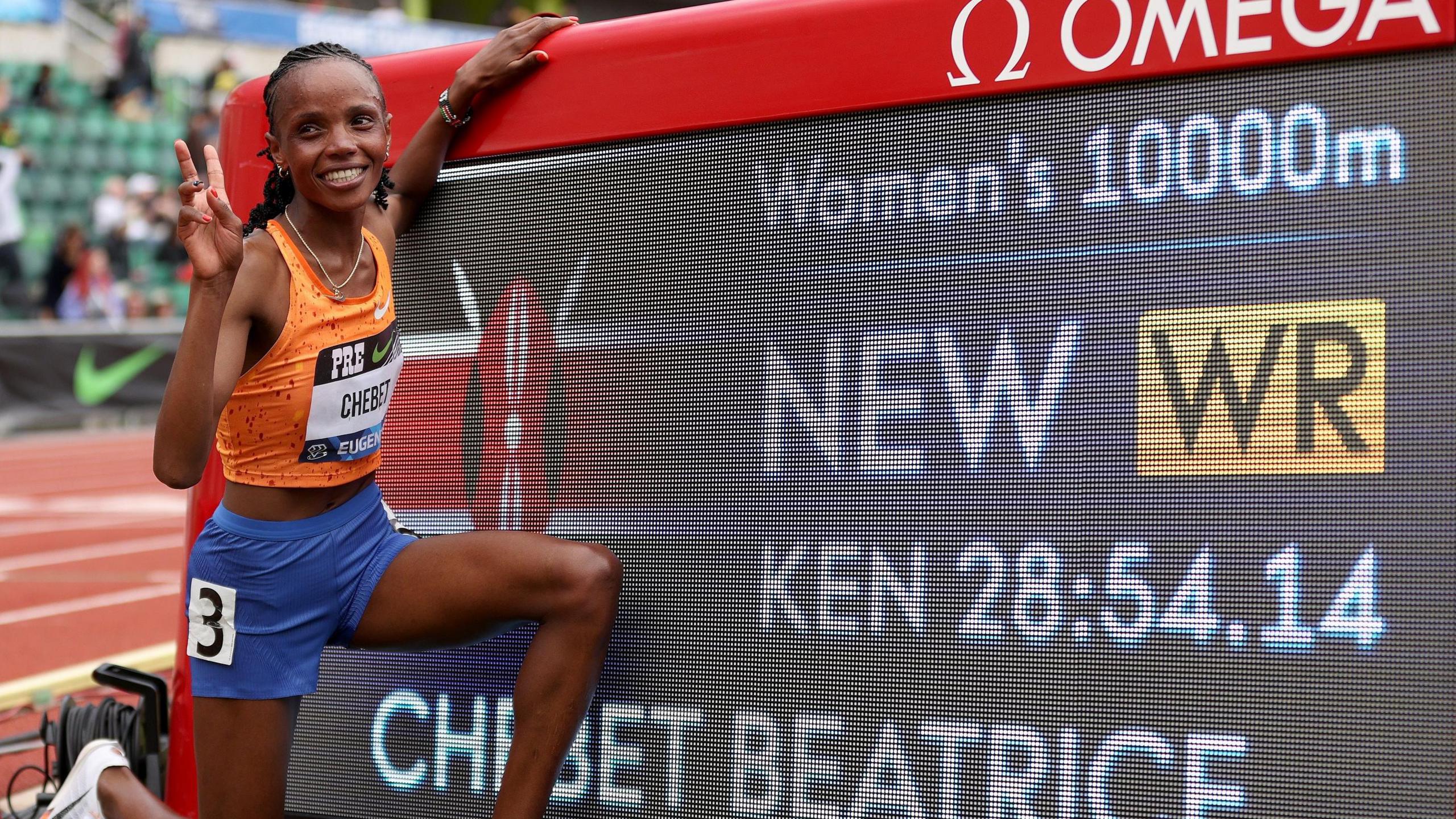 A smiling Beatrice Chebet, wearing an orange running vest and blue shorts, kneels down in front of an electronic scoreboard which displays a new world record time for the 10,000m of 28 minutes and 54.14 seconds. She has one hand resting on the scoreboard while with the other she makes a V sign. A running track is seen out of focus in the distance behind her