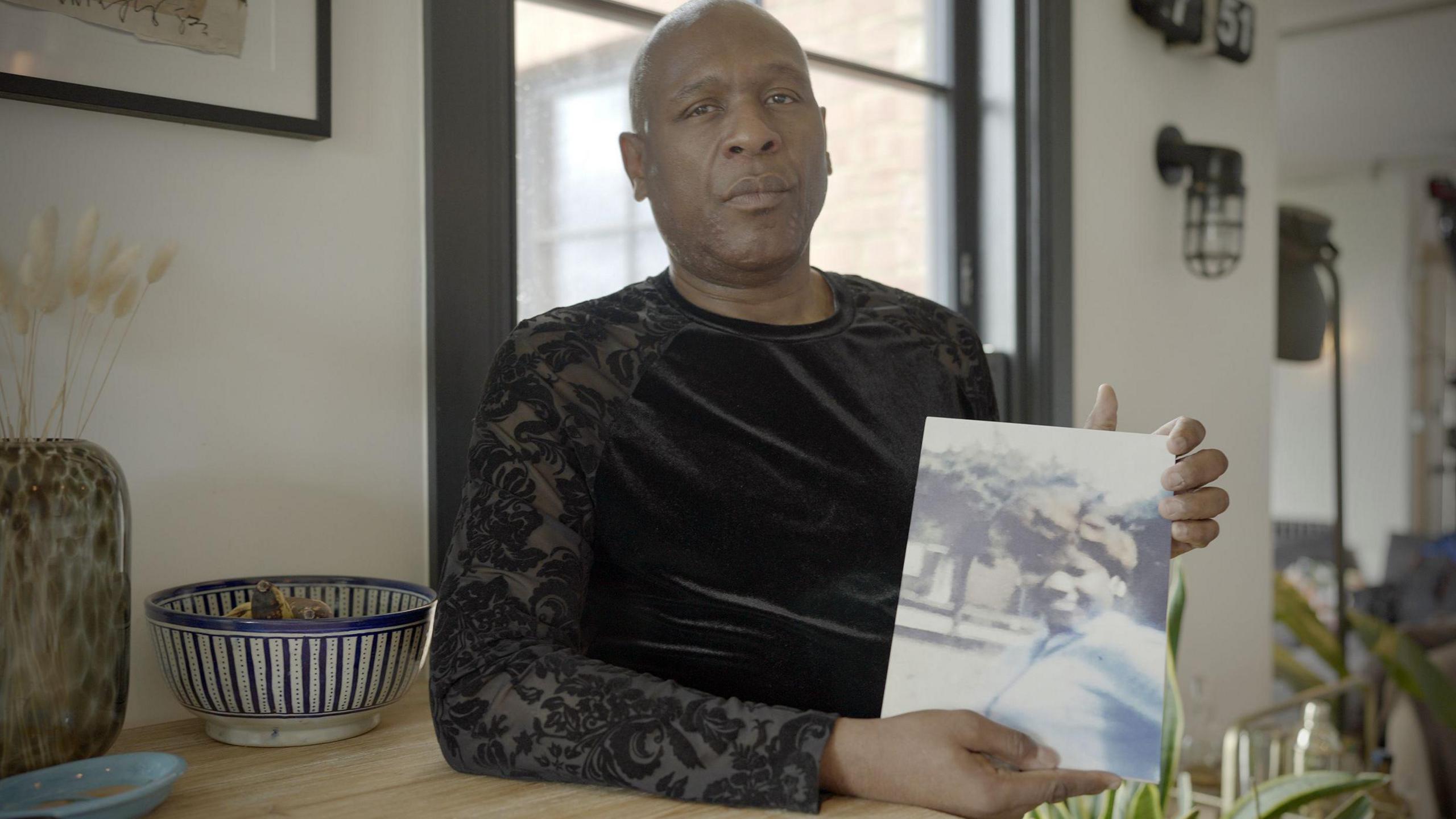Colyn Gordon, a bald man in a black patterned T-shirt, is holding a photo of his sister Olive and looking towards the camera. He is sitting at a pine table in a magnolia room.