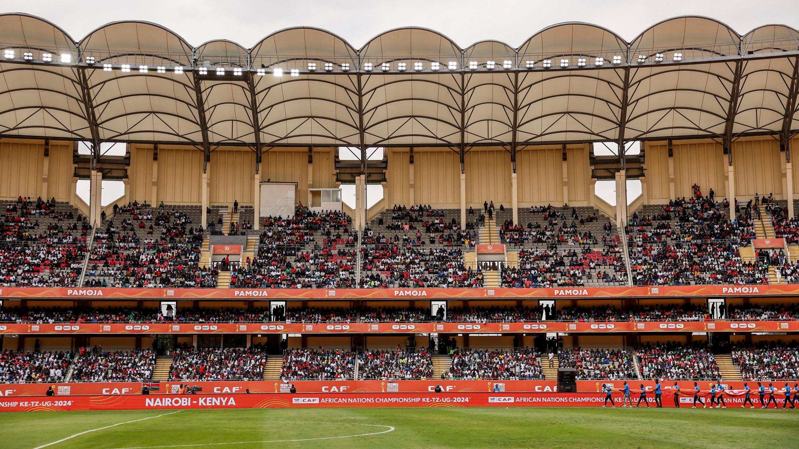 A wide view of the Kasarani Stadium, showing three tiers of packed stands and a canopy roof. The stands are decked in orange CHAN branding boards featuring the words Caf and Pamoja in white lettering, while part of a green football pitch can be seen in the foreground