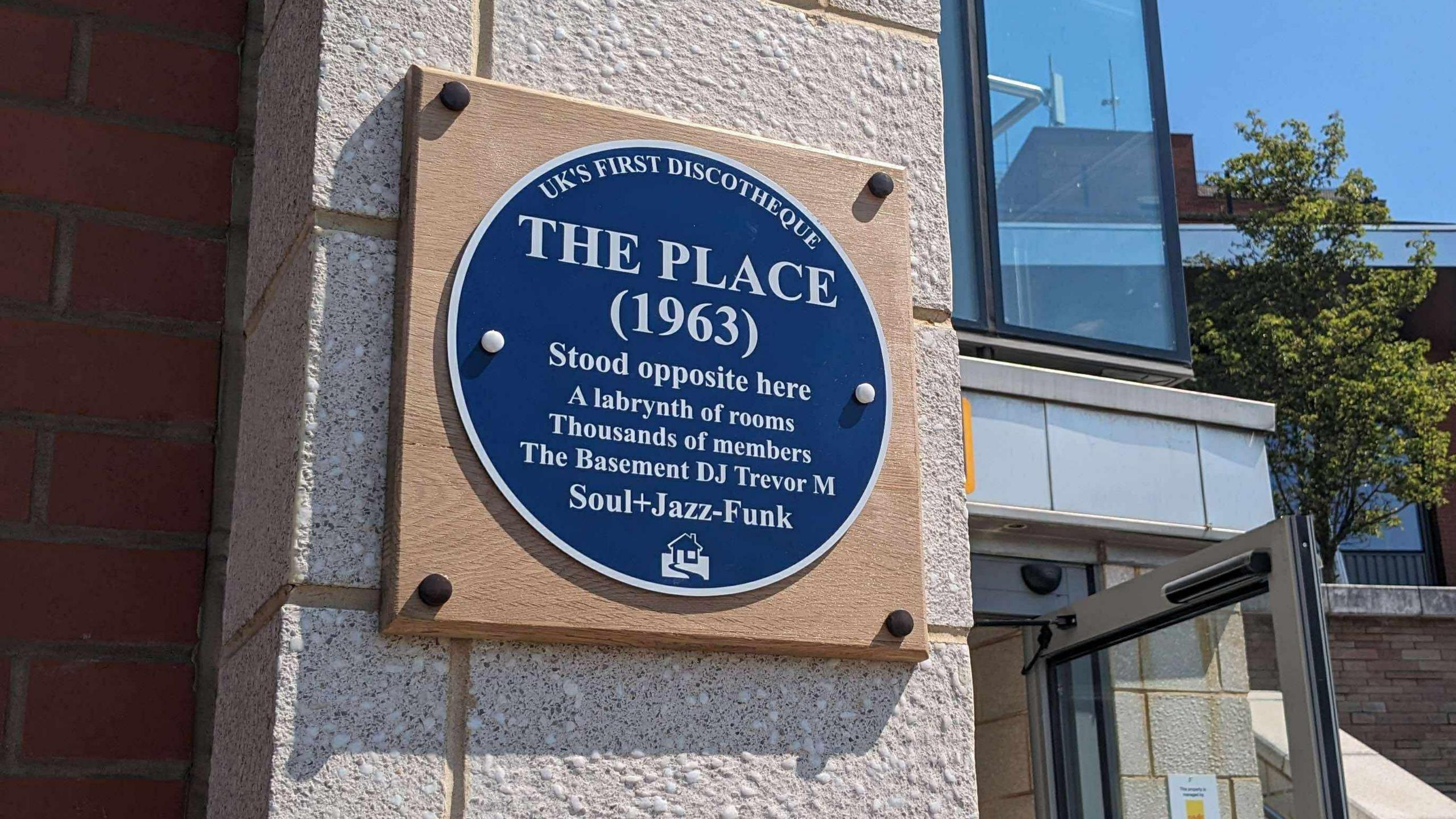 A blue plaque which is mounted onto a wooden square on the side of a building. The plaque has white writing which is titled "The Place" and details the site's history.