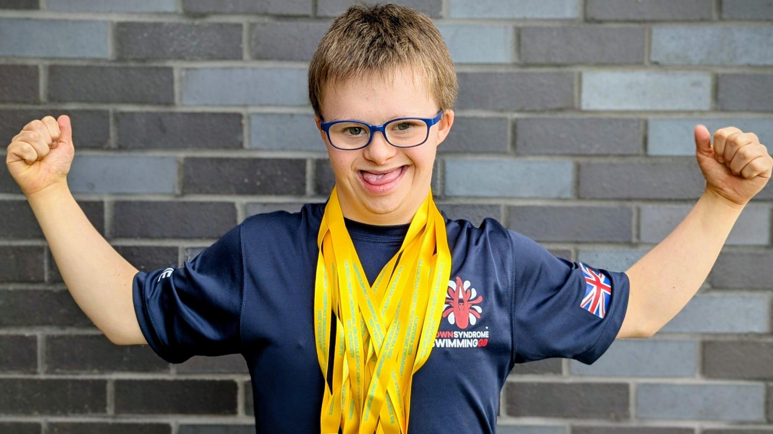 Joe Hughes, with two arms stretched out, with lots of yellow ribbons round his neck, he is smiling. He has on blue glasses and has short fair hair. He is standing by a brick wall and has on a blue T-shirt. 