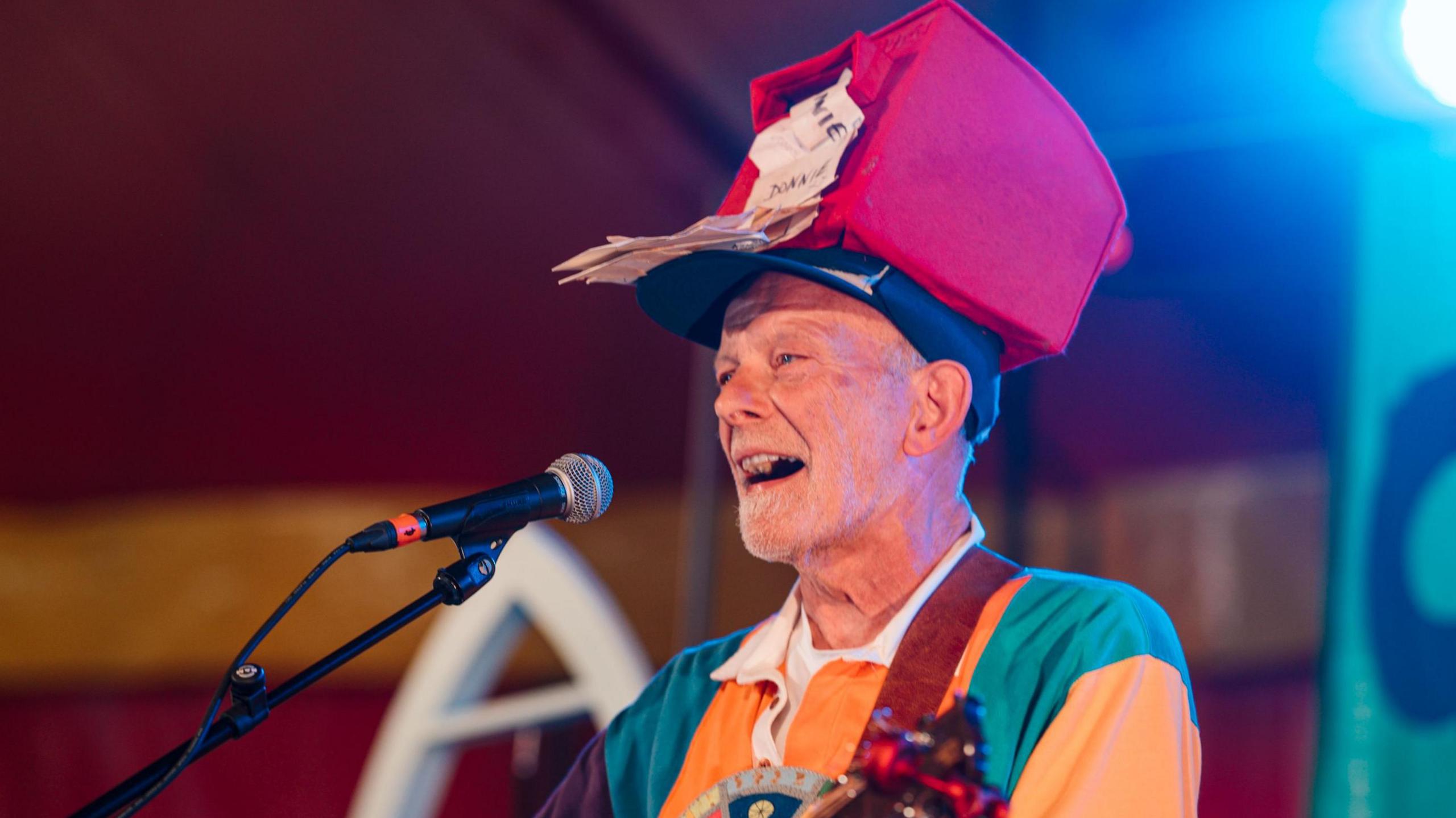 Donnie Dòtaman is singing or speaking into a mic. He is wearing a red hat made to look like a post box with letters coming out of it. He is wearing a multi-coloured top