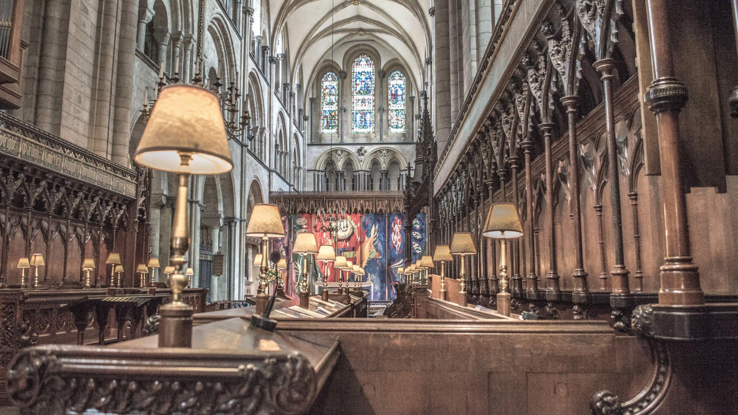 The choirstalls inside Chichester Cathedral, with a large stained glass window in the background.