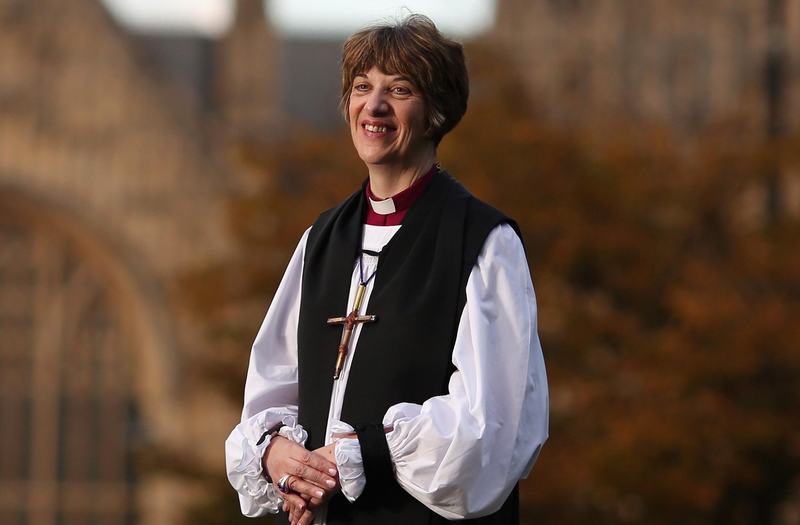 The Bishop of Gloucester Rachel Treweek stands in her full clergy outfit including a large crucifix. In the background are trees and a church building