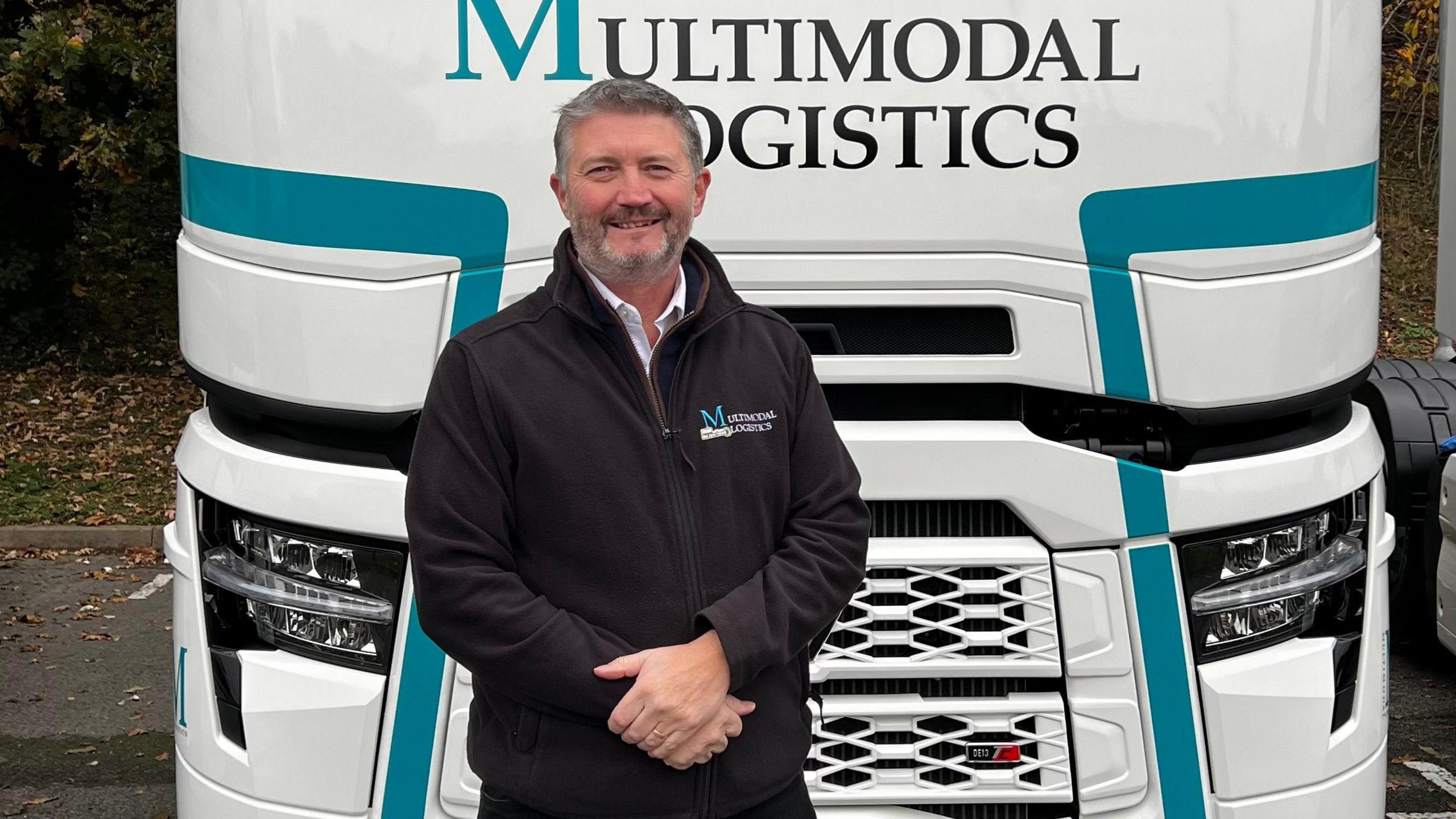 Tim Wray smiles at the camera in front of a white HGV lorry. He has short grey hair and wears a black jumper with his hands resting in one another in front of him.