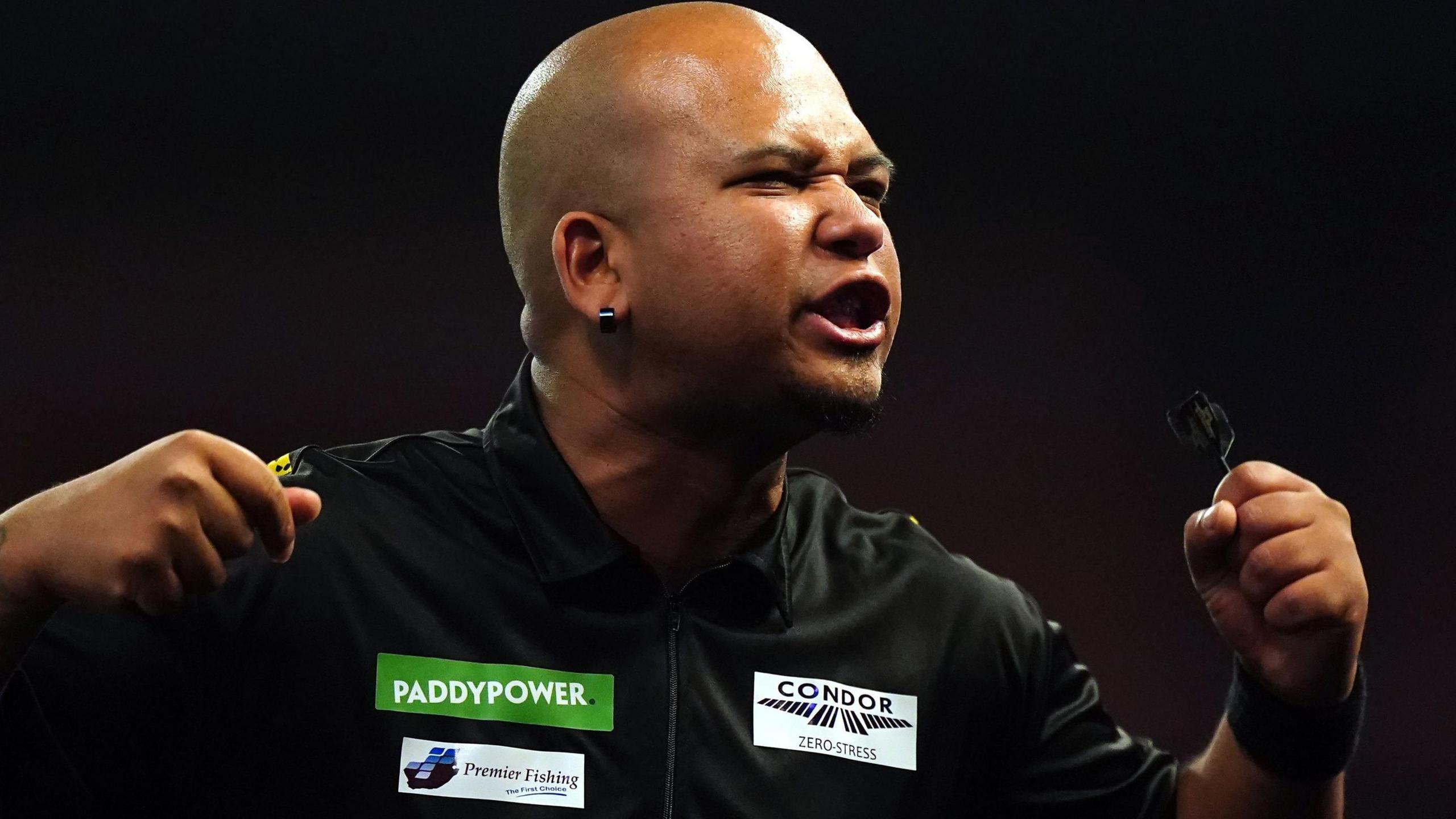 Cameron Carolissen, holding a dart in each hand with fists clenched, grimaces and roars at a crowd which is off camera