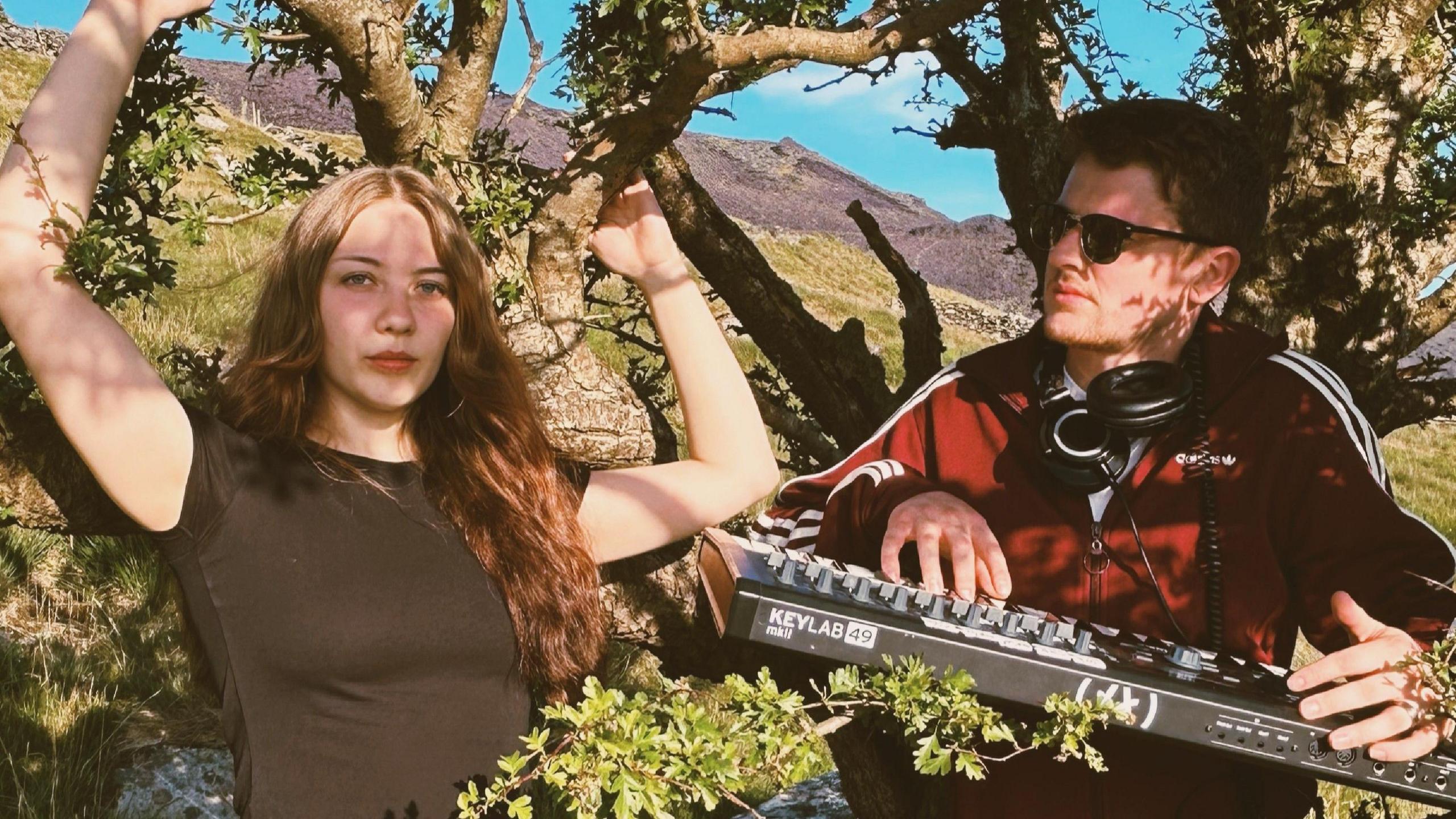 Ruby ar y chwith ac Endaf ar y dde yn sefyll mas tu fas wrth ymyl coeden. Mae gan Ruby wallt hir brown ac mae'n gwisgo crys-t du. Mae Endaf ar y dde yn gwisgo yn gwisgo clust-ffonau ac yn dal bysellfwrdd