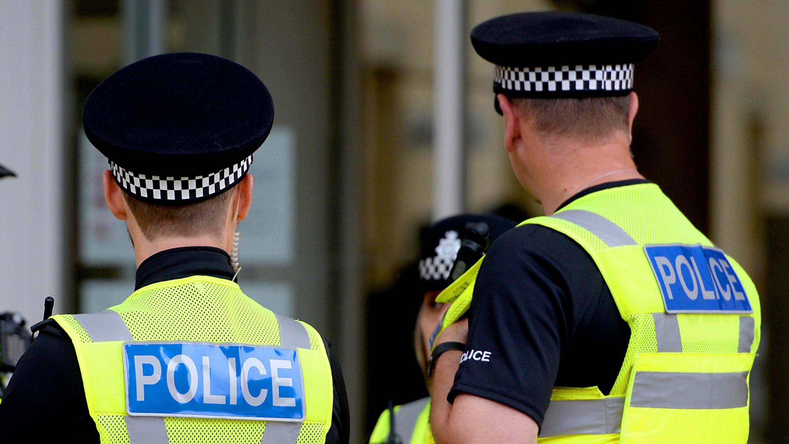 Two police officers with their backs to the camera, wearing police caps and two hi-vis yellow jackets with the signs reading police, in blue, on their backs. 