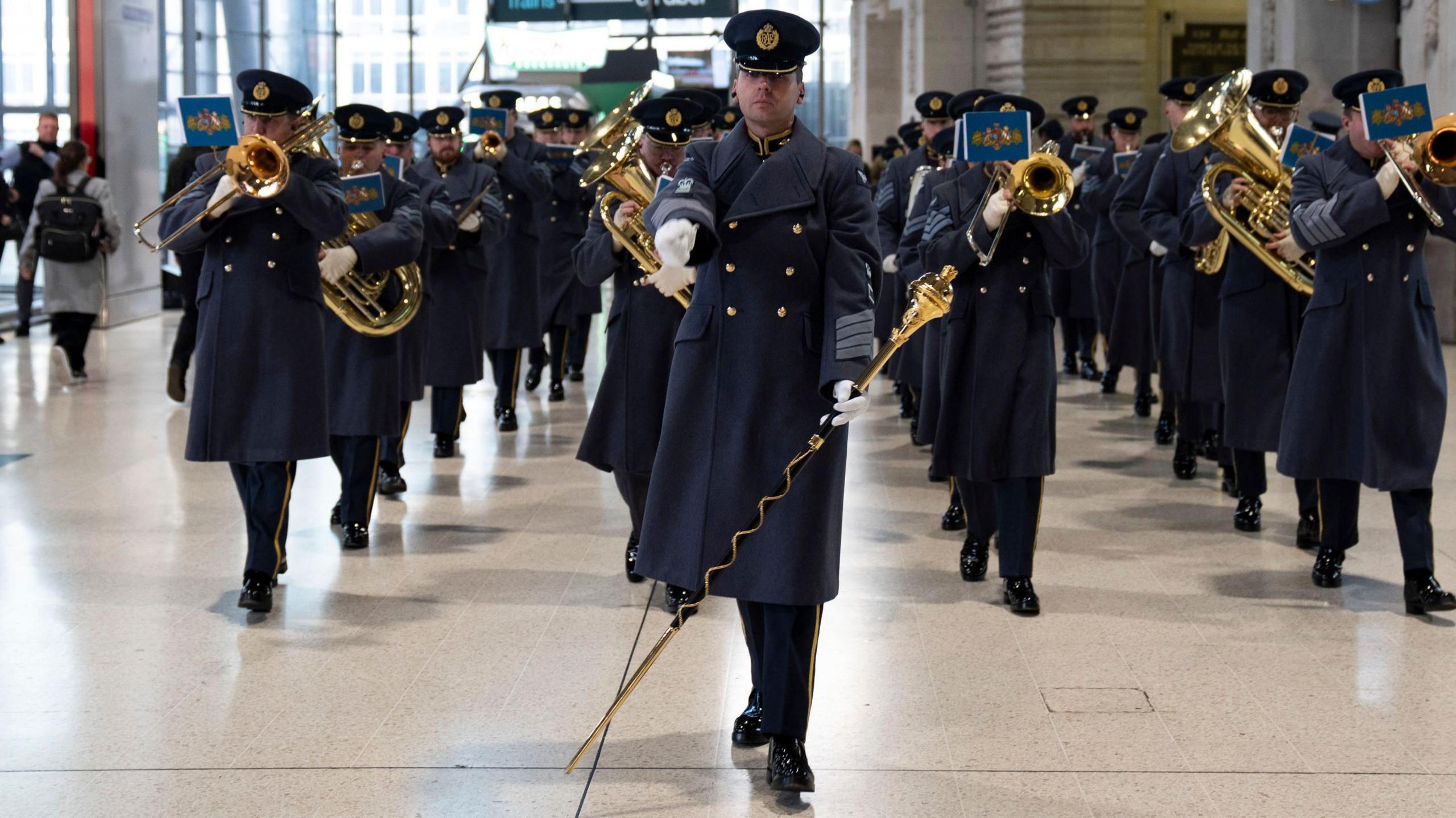 An RAF band at the launch of the Royal British Legion's annual London Poppy Day, at Waterloo Station in London. They are dressed in blueish grey overcoats and hats and many of them are playing brass instruments.