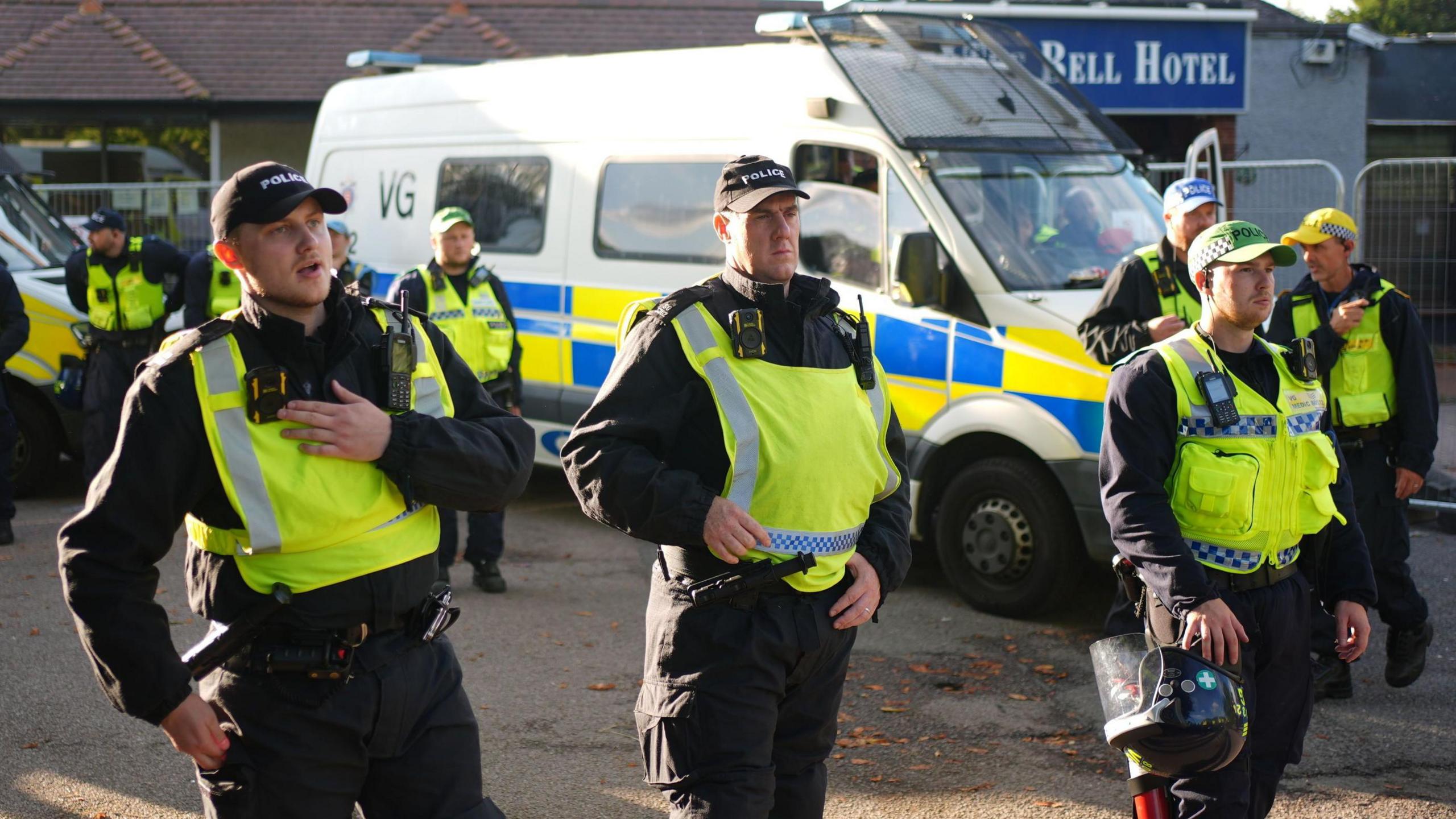 A group of police officers in fluorescent tabards speaking into talkback devices and holding riot helmets - there is a large police van in the background at the entrance to a building with a Bell Hotel sign 
