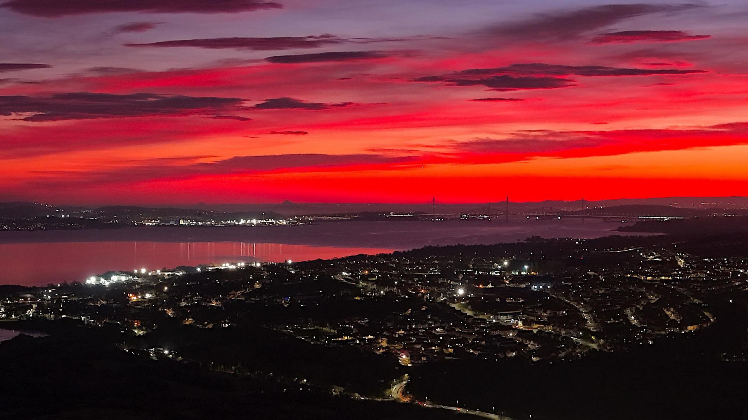 Brilliant shades of red and orange in the sky above West Lothian and Edinburgh in a picture taken at sunrise from atop a hill with the lights of the town of Bo'ness in the foreground. The outline of the bridges over the Firth of Forth can also be made out. 