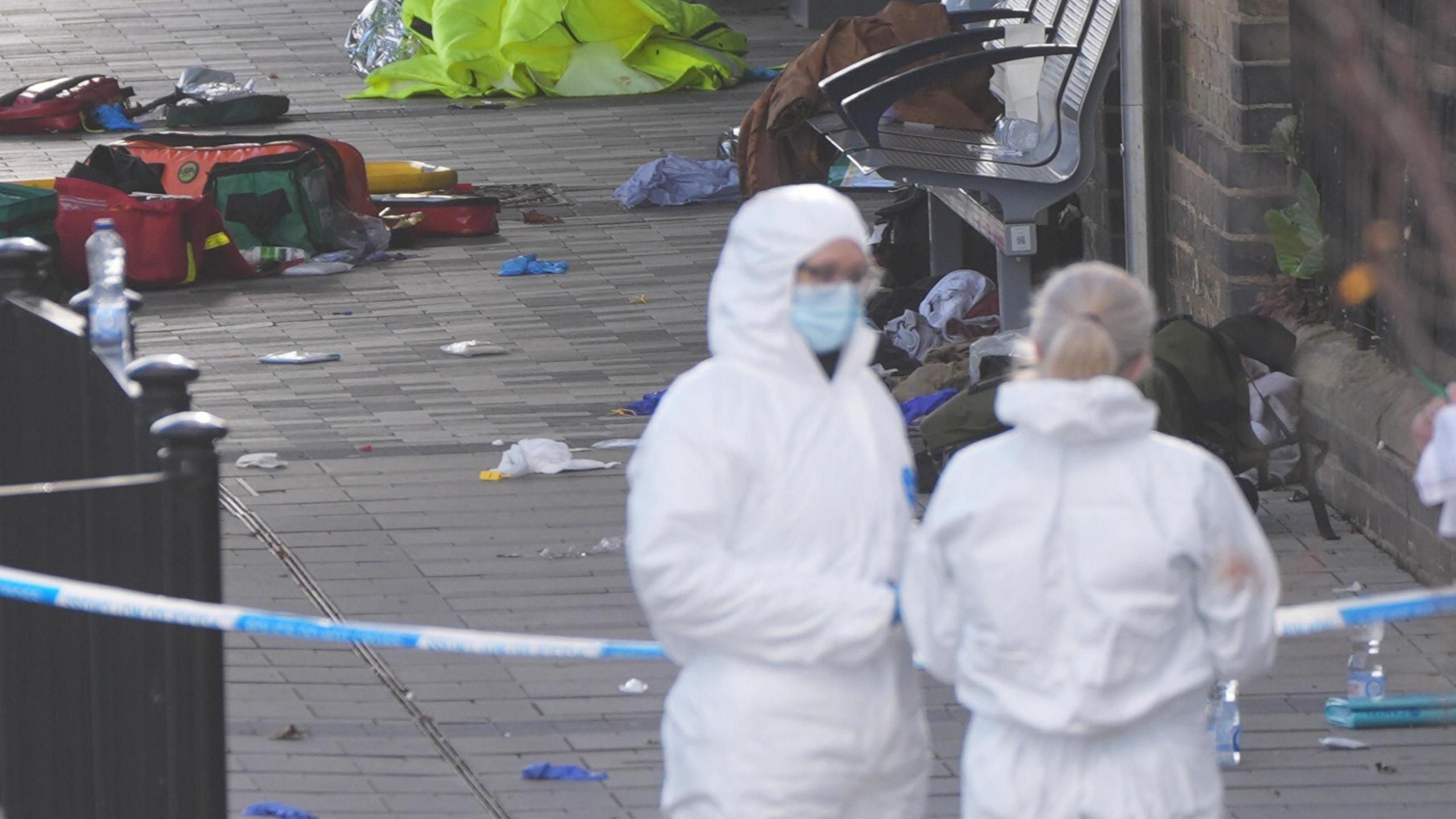 Two people are in white overall suits standing on a train platform. There is rubbish on the floor and paramedic bags. 