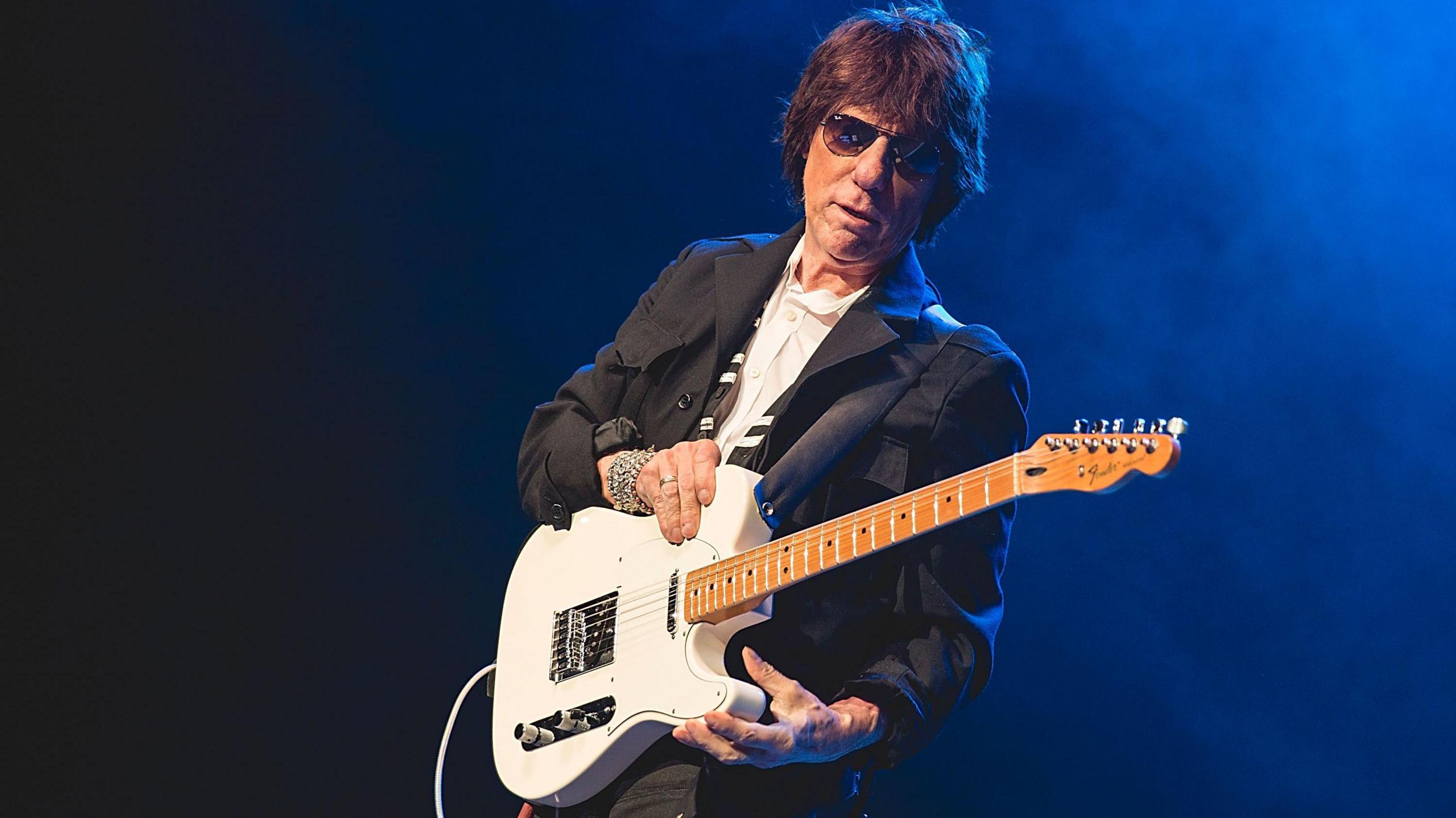 Jeff Beck performing on stage, wearing a white shirt, black jacket and trousers and sunglasses, playing a white Fender Stratocaster guitar.