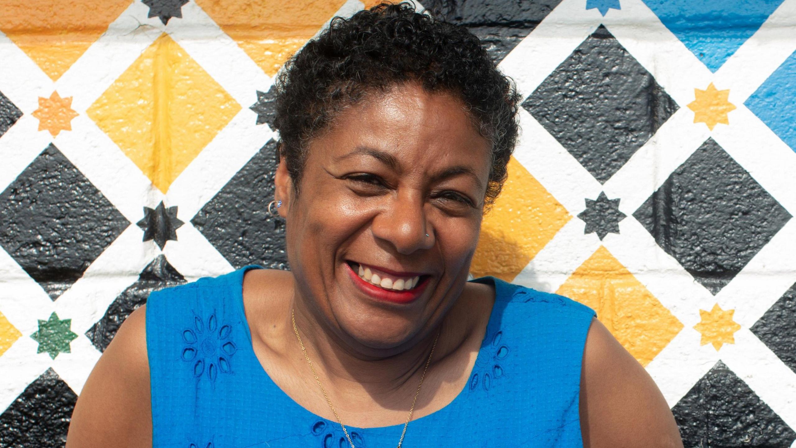 Patrice Lawrence wearing a blue dress and smiling at the camera. She is sitting in front of a colourful wall.