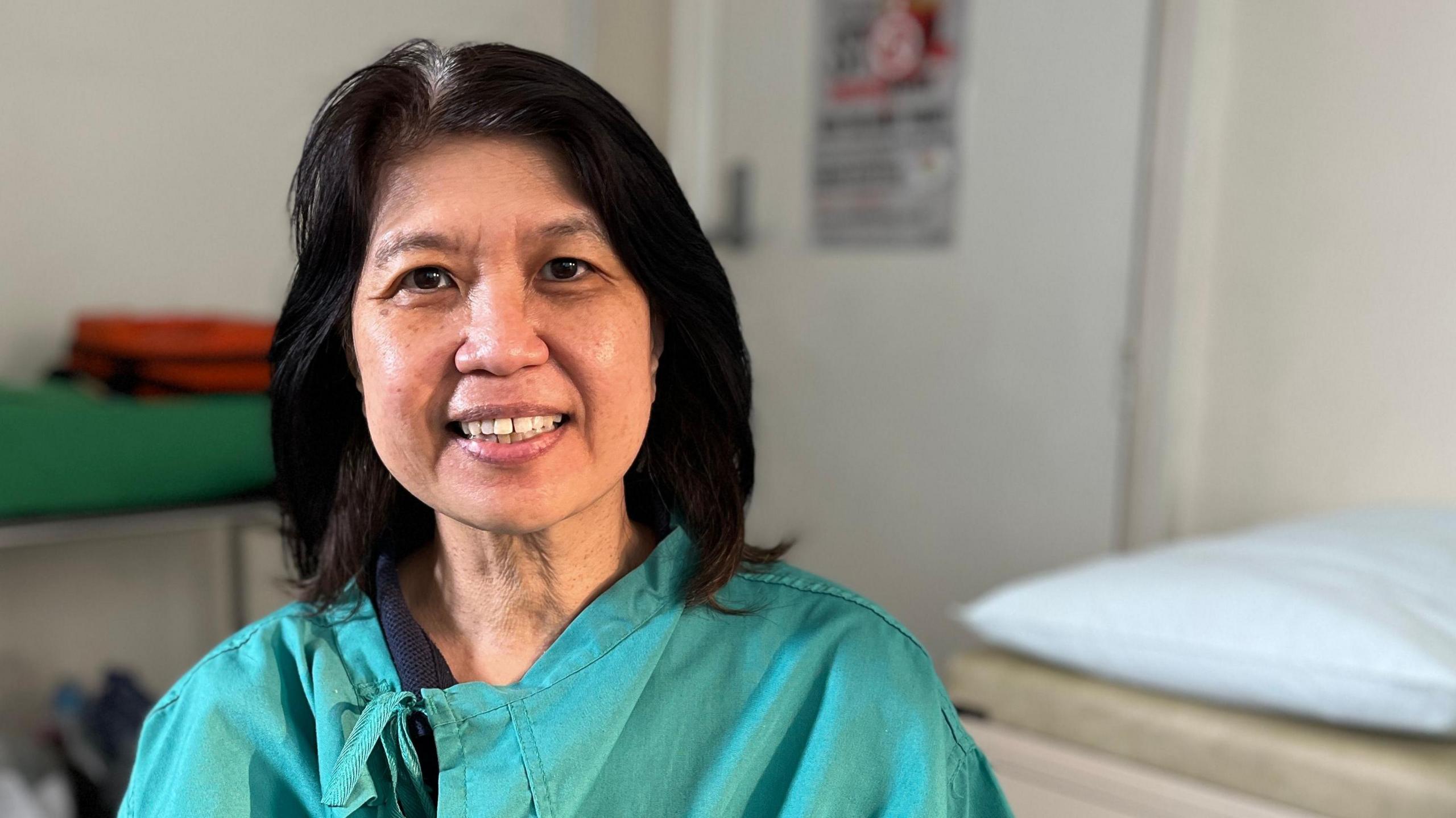 Jennifer Loke sits in a doctor's consulting room with an examination couch behind her. She is smiling, has long black hair and is wearing green scrubs.