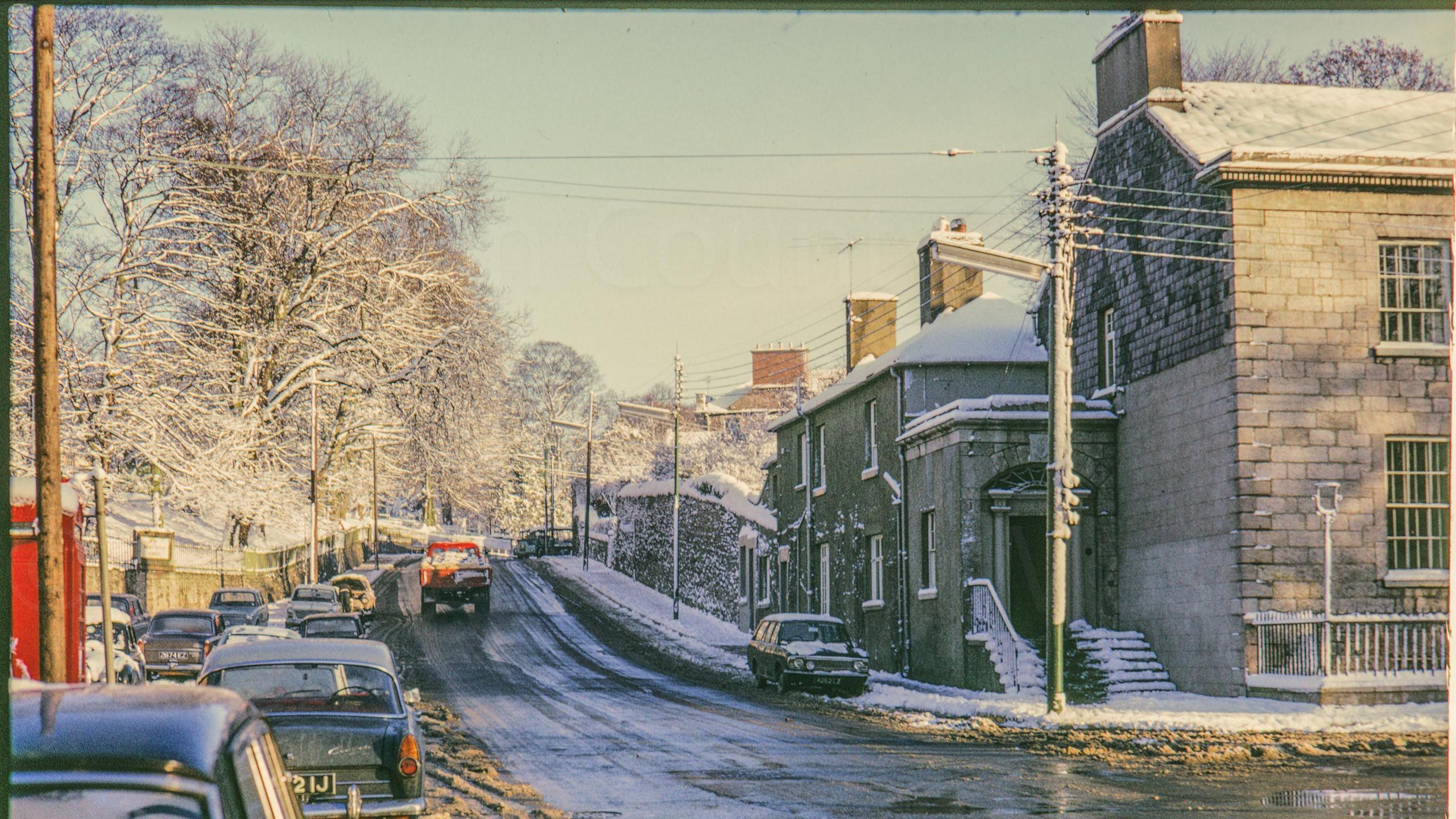 A digitalised photo from a film of a snowy street. Large stone buildings are along the right and trees and cars line the left. College Hill Armagh in snow from junction with Mall East showing Royal Irish Fusilier's museum (1 Beresford Row) on right.