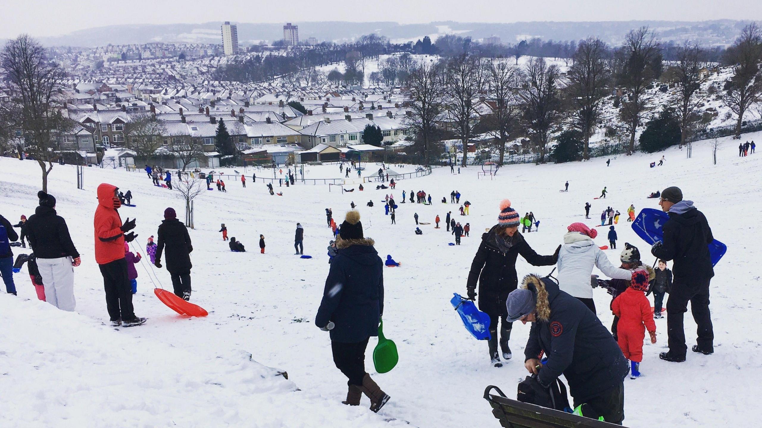 A view looking down on Perrett's Park in Bristol during heavy snowfall during the so-called "Beast from the East" storm in 2018. Many people are visible in winter clothes riding sledges down the steep slope, and the snow-covered rooftops of Totterdown and Victoria Park are visible in the background