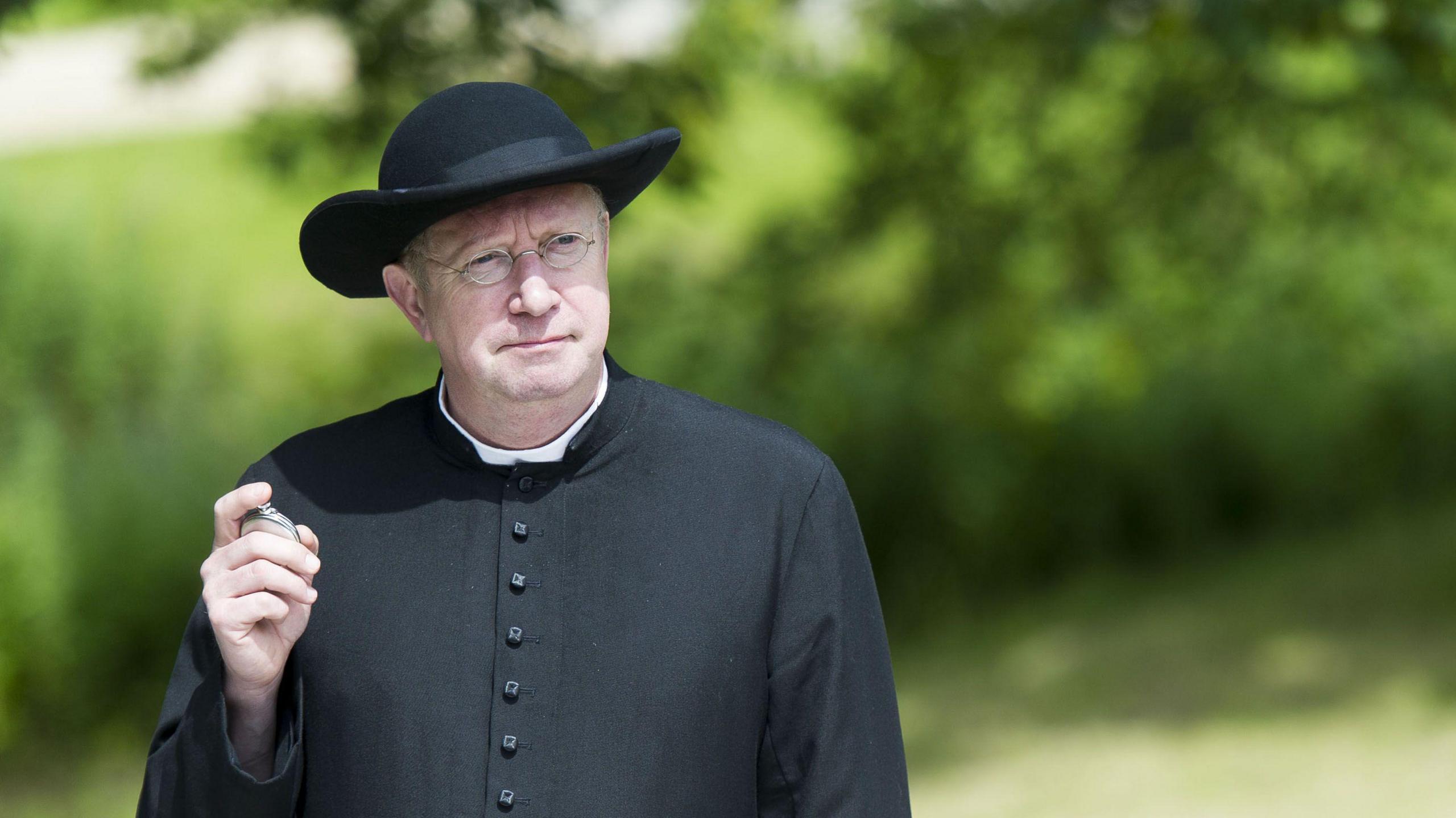 Mark Williams as Father Brown. He is dressed in black clerical robes and a wide-brim hat. He is holding a silver pocked watch and is also wearing thin, silver glasses. 