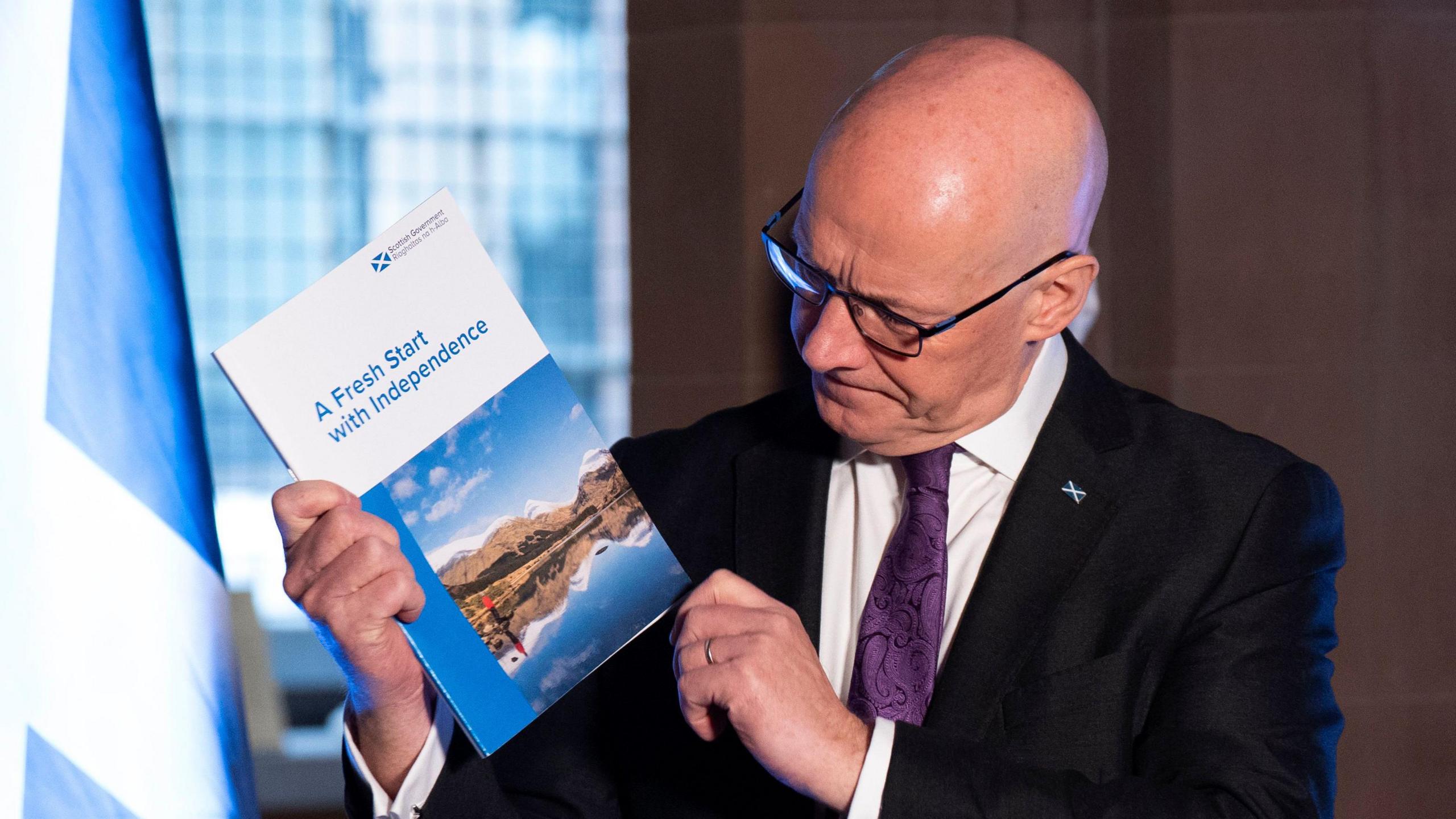 John Swinney - in the same outfit as the previous photo - stands holding the glossy booklet and looking at the front of it which says: "A fresh start to independence". There is a window and a Saltire in the background