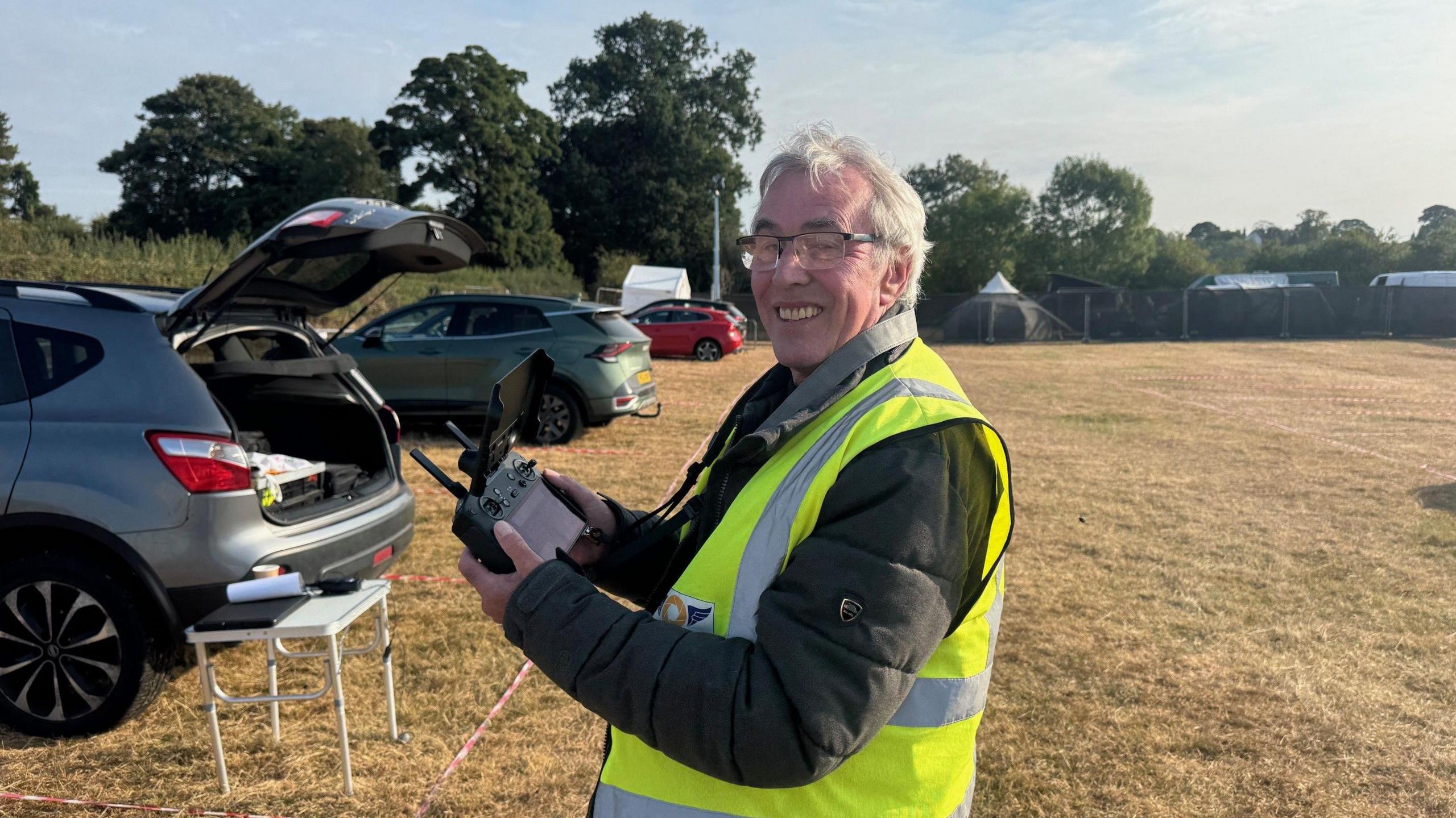 An older man with short, grey hair and wearing a yellow hi-vis jacket is standing in a field. The grass is dry and he is holding a drone controller. He is smiling and standing side on with his head turned towards the camera.  There are a few cars parked in front of where he is standing.