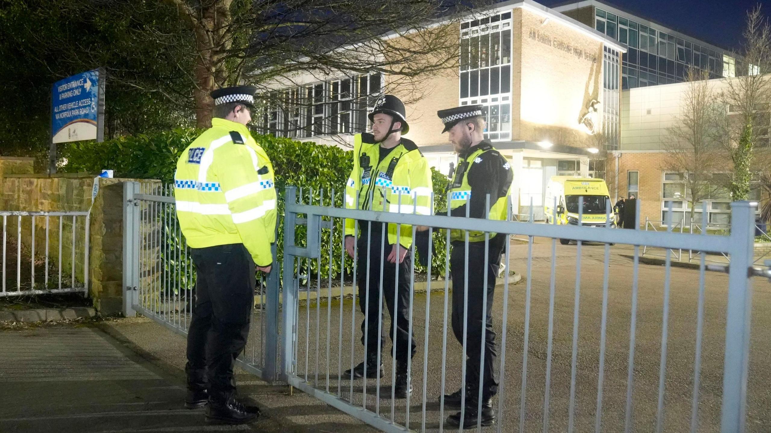 Three police officers stand by a gate in front of a school building