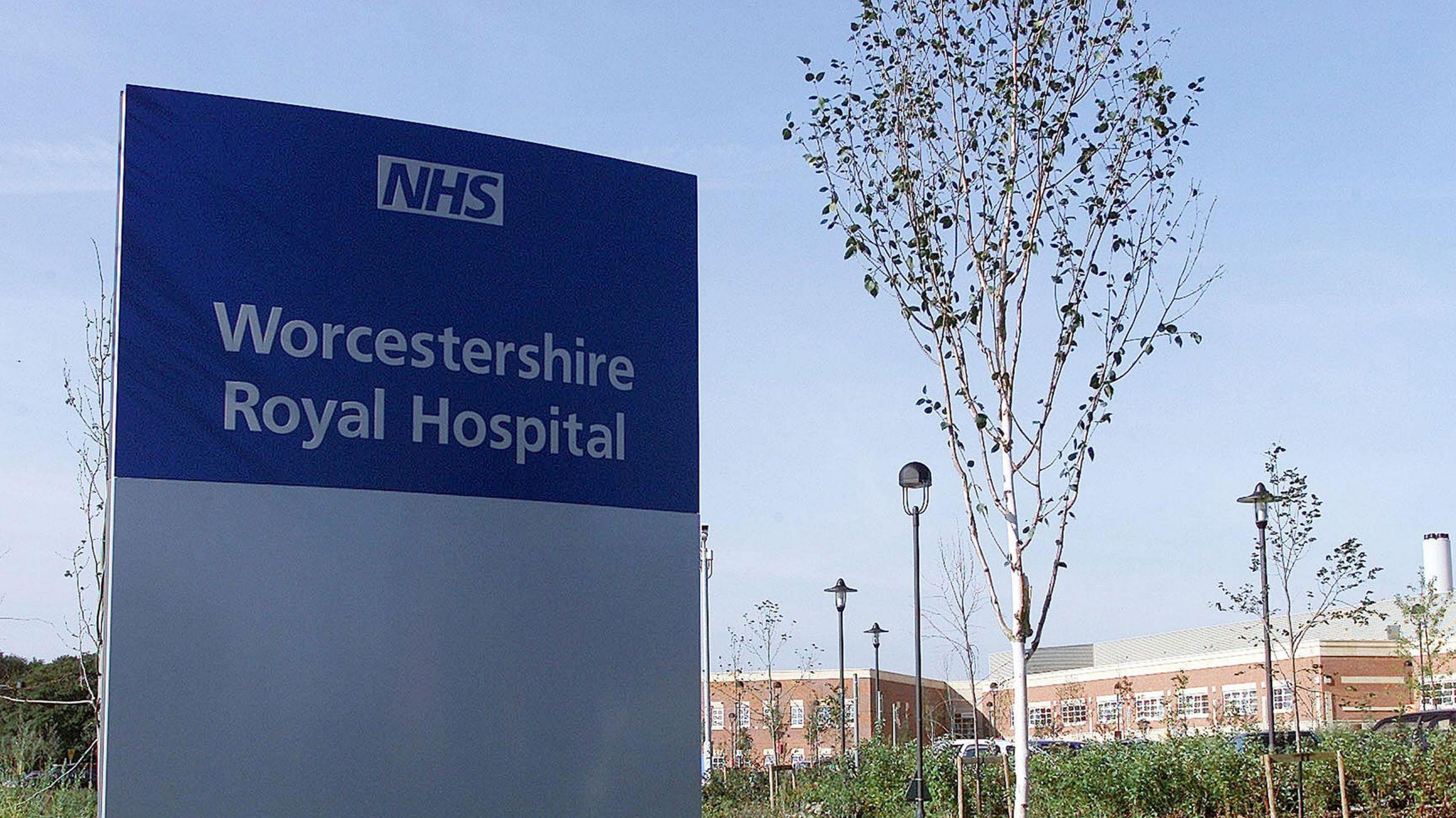 A rectangle-shaped sign that has a silver base with a blue top. The sign reads "NHS Worcestershire Royal Hospital". There are trees and a red brick building behind the sign.