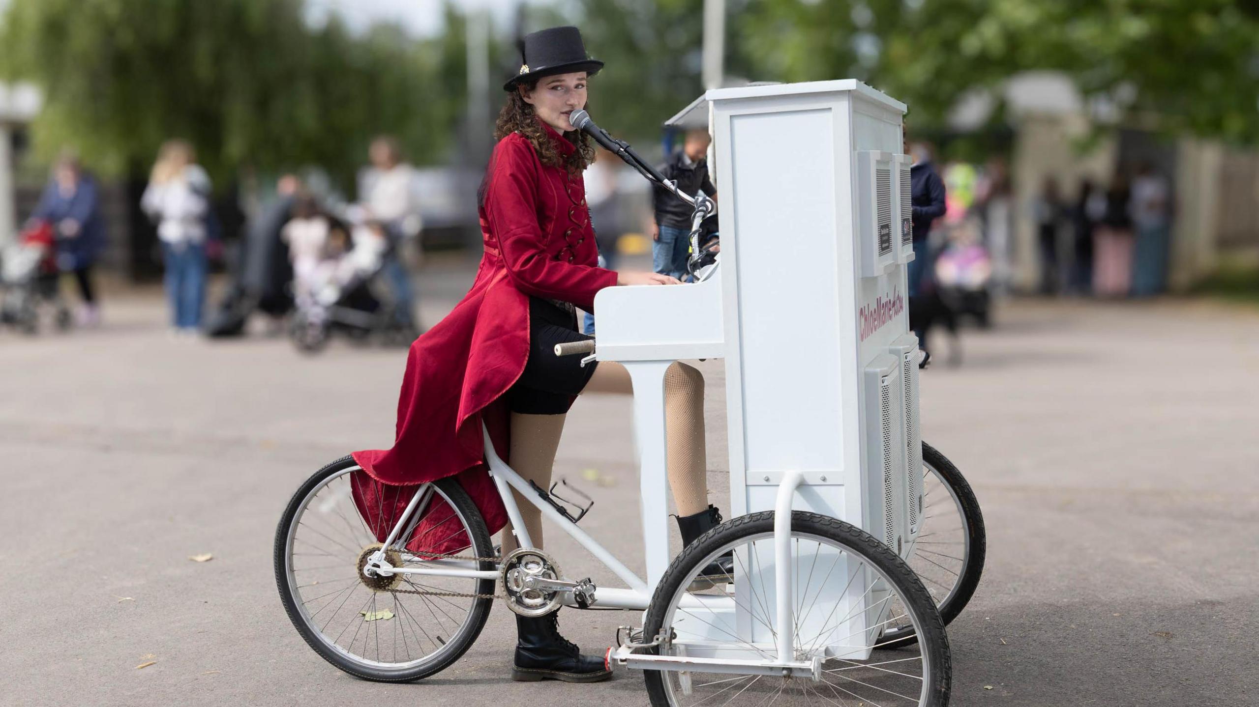 A woman performer at the Bath and West Show sings into a microphone coming out of a white piano which is balanced on the front of a tricycle, where the handlebars would normally be. She is wearing a long, red coat and a black top hat.