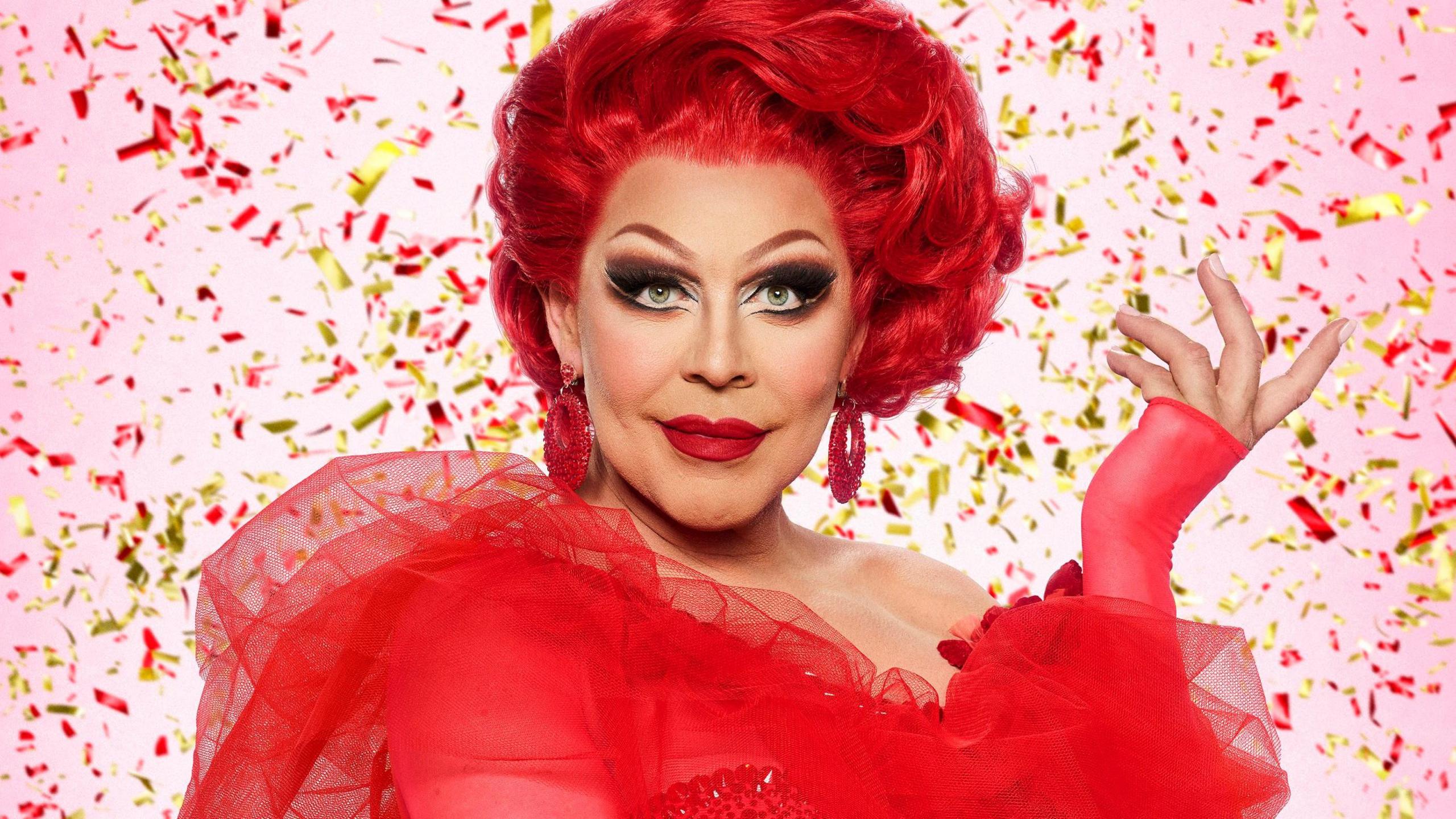 La Voix, with a thick red mane of hair and wearing a red dress, stands against a pink background surrounded by gold and red confetti