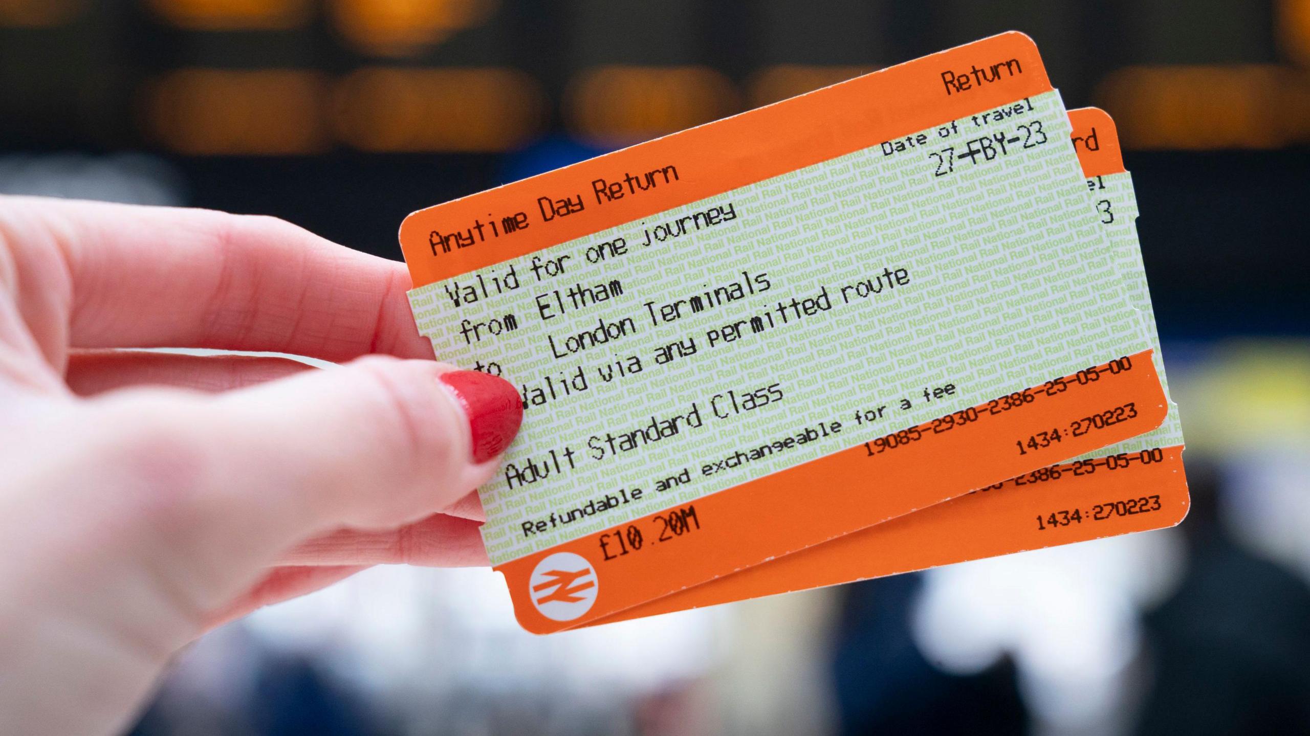 A woman with red nail varnish holding a rail ticket to from Eltham to London terminals in front of a departure board