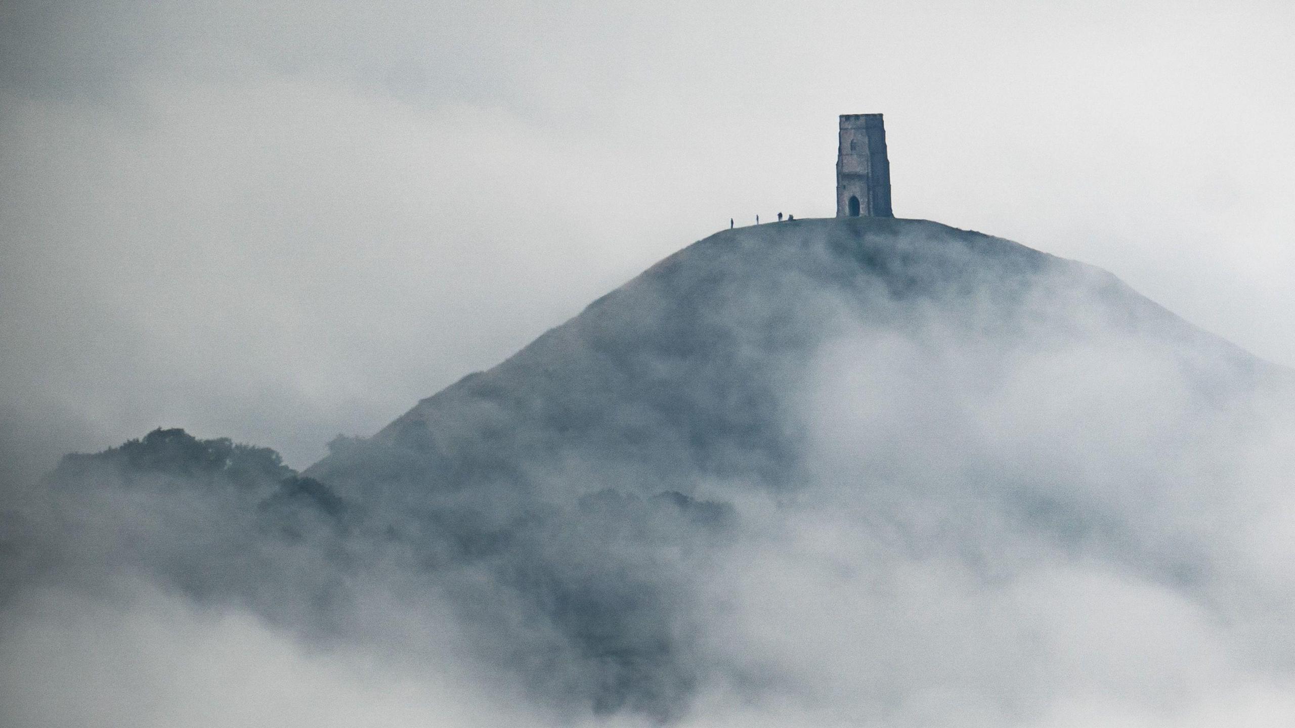 St Michael's Tower on Glastonbury Tor. Small silhouettes of people can be seen on top of the hill. There is lots of mist and grey clouds in the picture. 