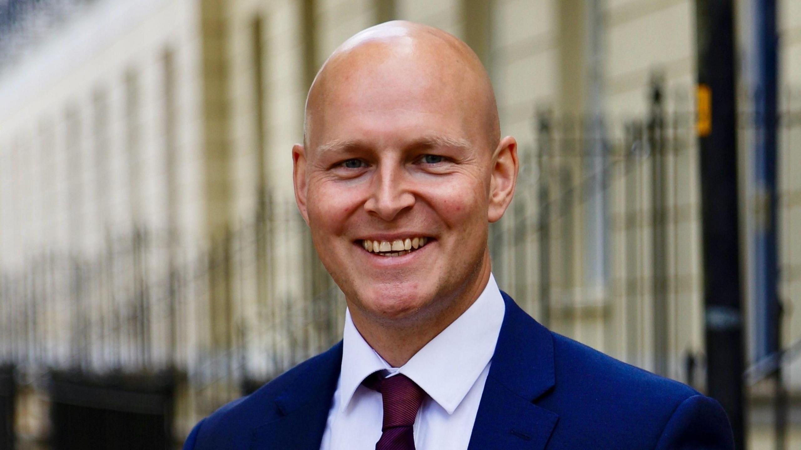 Max Wilkinson wearing a red tie, white shirt and blue blazer. He is standing in the street, smiling at the camera. 