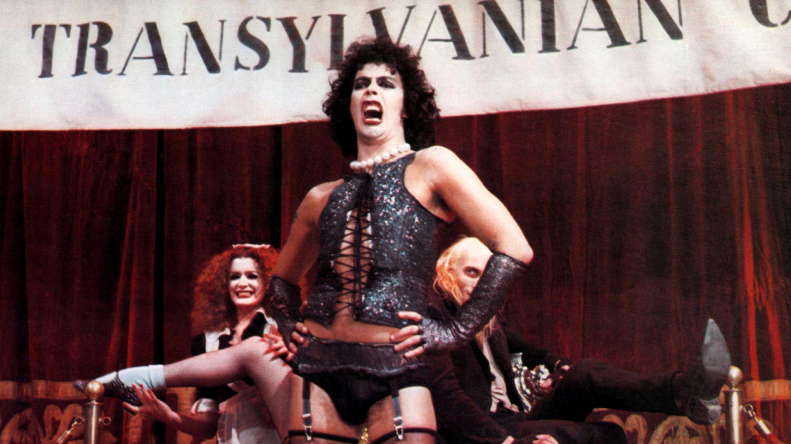 Tim Curry in a scene from the Rocky Horror Picture Show, wearing a black corset and stockings. He is singing in front of a Transylvania banner as other cast members watch