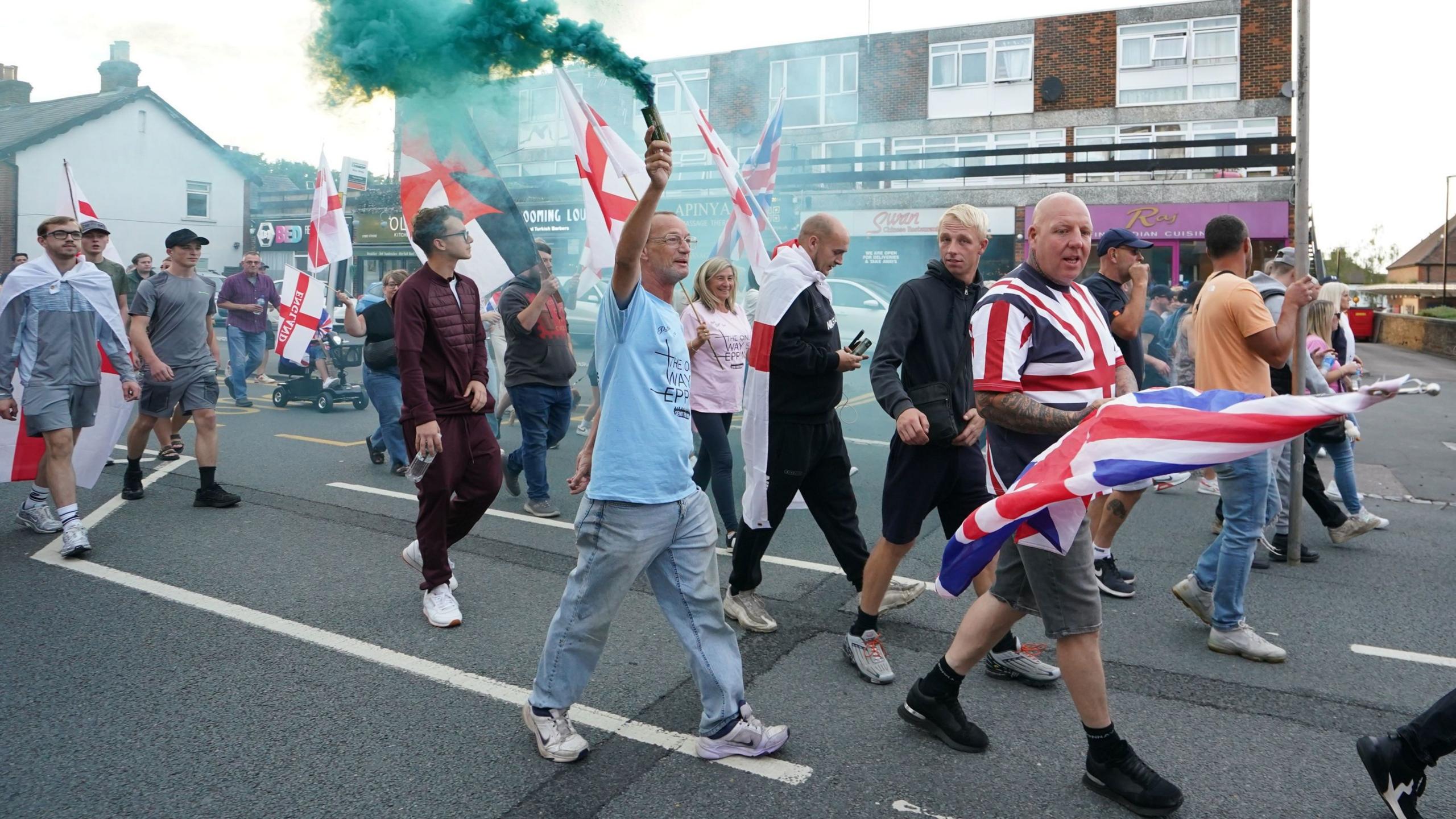 Protesters walking along a road wearing and carrying union jacks and St George cross flags with the man in the centre of the picture carrying a green smoke grenade