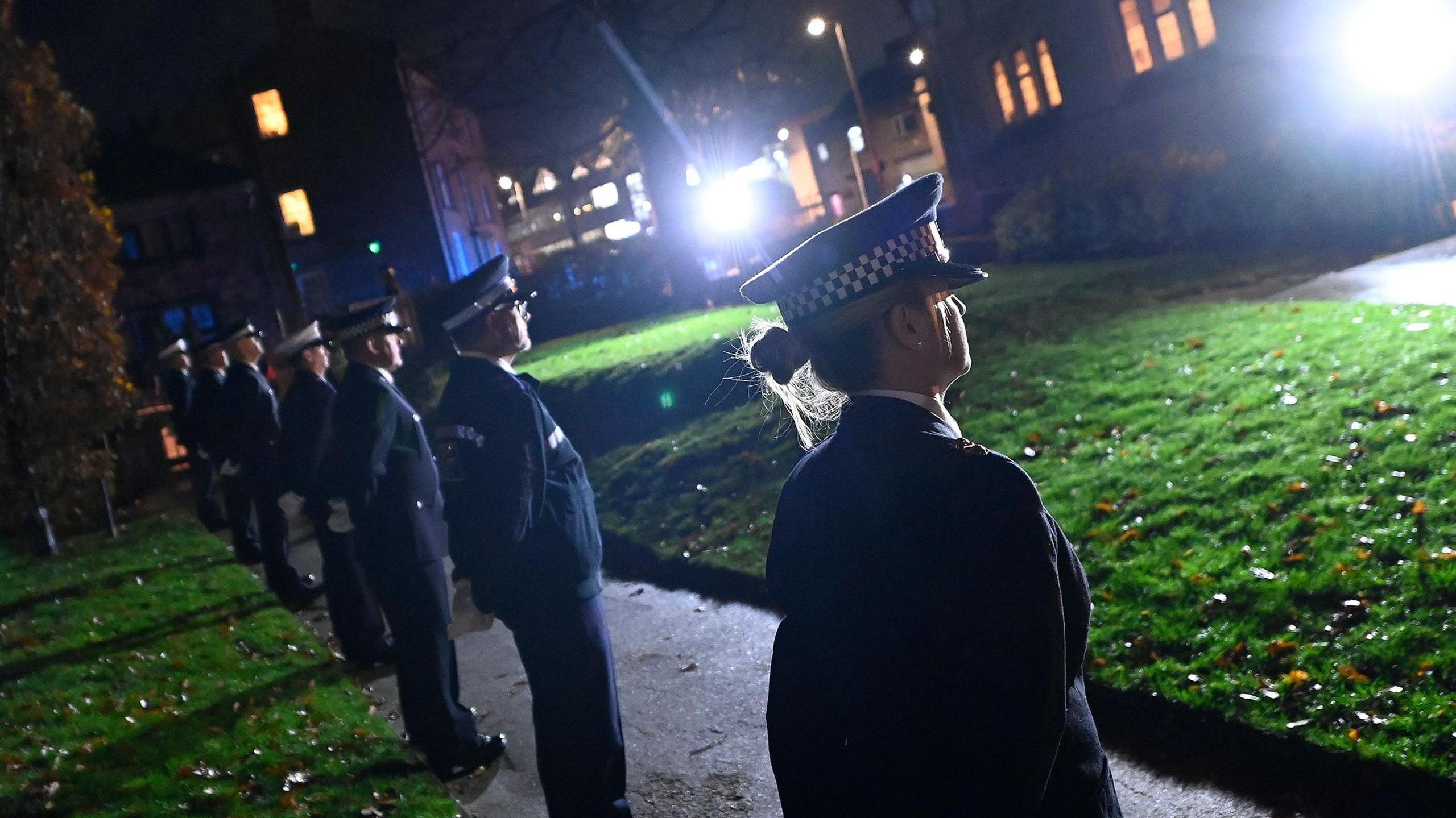 A line of people wearing uniforms and caps standing in a row with their hands behind their backs on a pathway near a grassed area. The scene is outdoors at night.