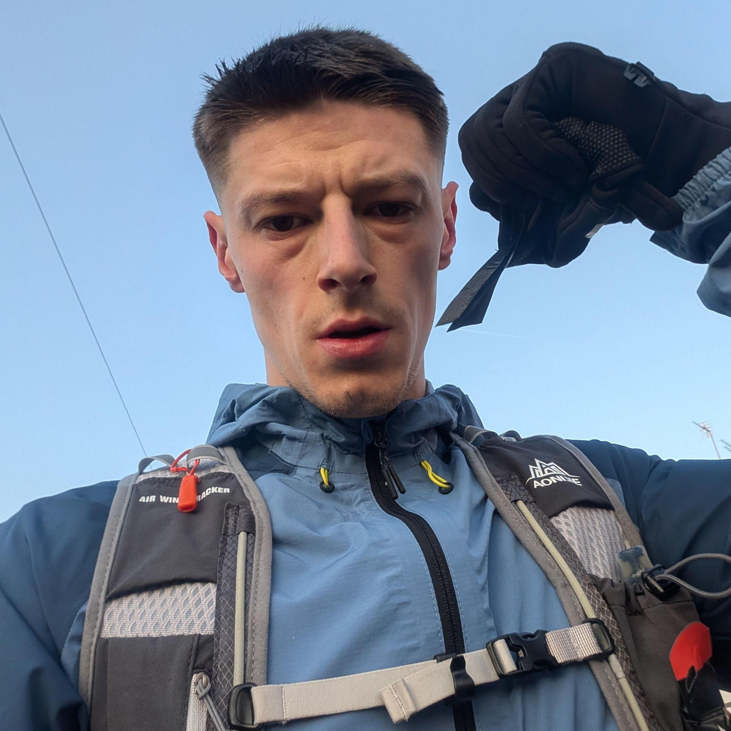 A man with short hair wearing a blue coat, black gloves and a grey rucksack takes a selfie while raising his left hand.