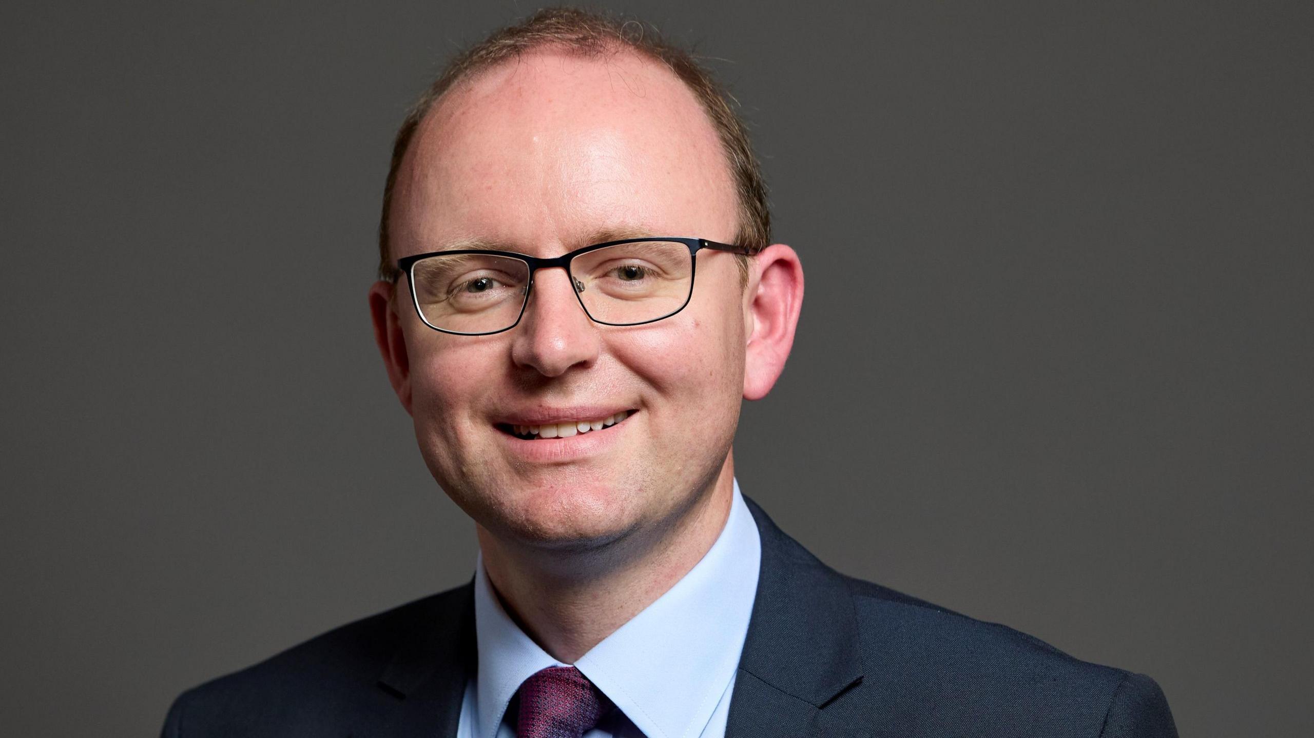 Andrew Cooper, the MP for Mid Cheshire, has receding brown hair and wears glasses. He smiles for the camera while wearing a dark suit, pale blue shirt and purple tie.