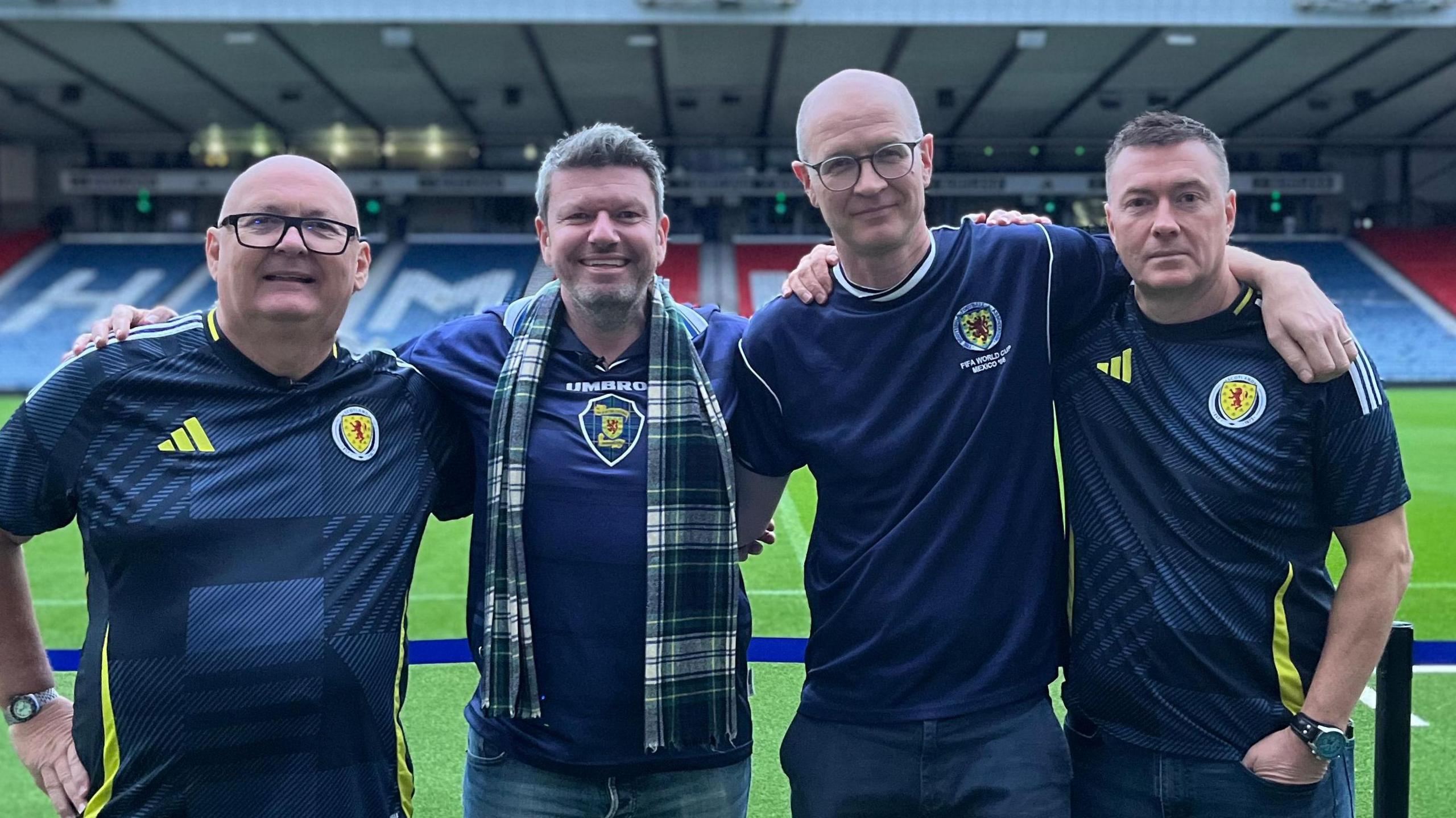 Four male Scotland fans, all in navy blue Scotland jerseys, stand arm-in arm with the pitch and the seating stand behind them at Hampden Park