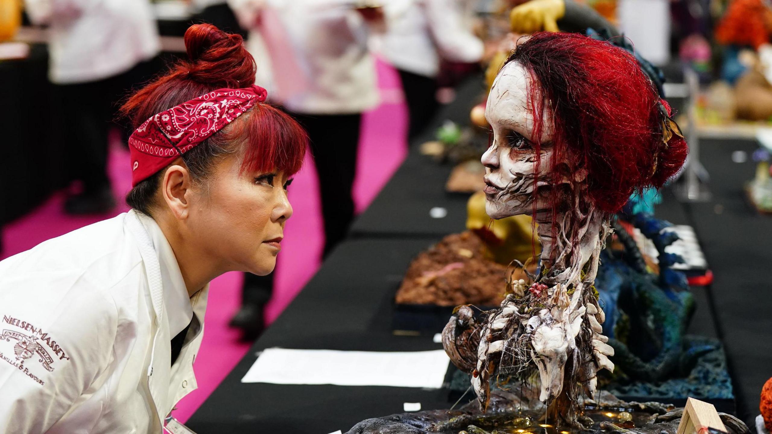 A woman with red hair, tied back with a red bandana, and wearing a white chef's top, closely inspects a cake depicting a disfigured woman's head and neck.