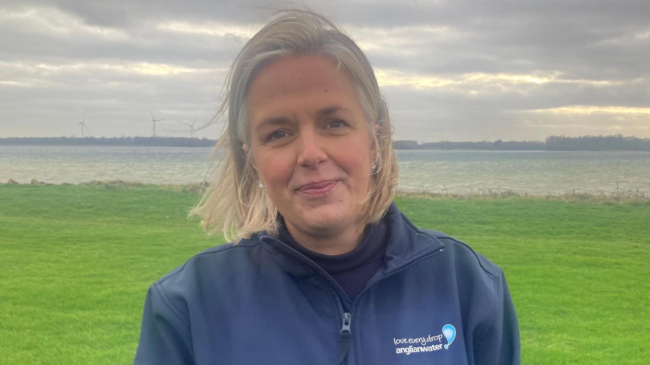 Emma Staples a woman with a short blonde and grey bob. She is wearing a navy zip up hoodie with an Anglian Water logo and slogan on. She is looking into the camera and smiling. In the background you can see a field and wind turbines.