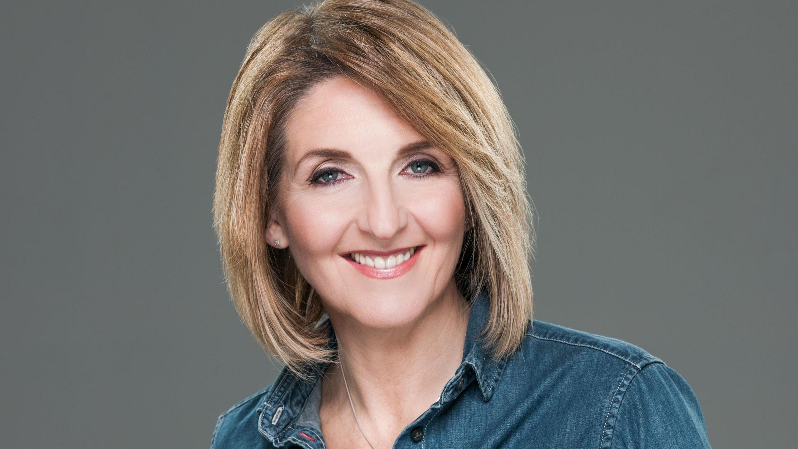 Kaye Adams, a woman with short blonde hair and wearing a denim shirt, looks into the camera smiling.