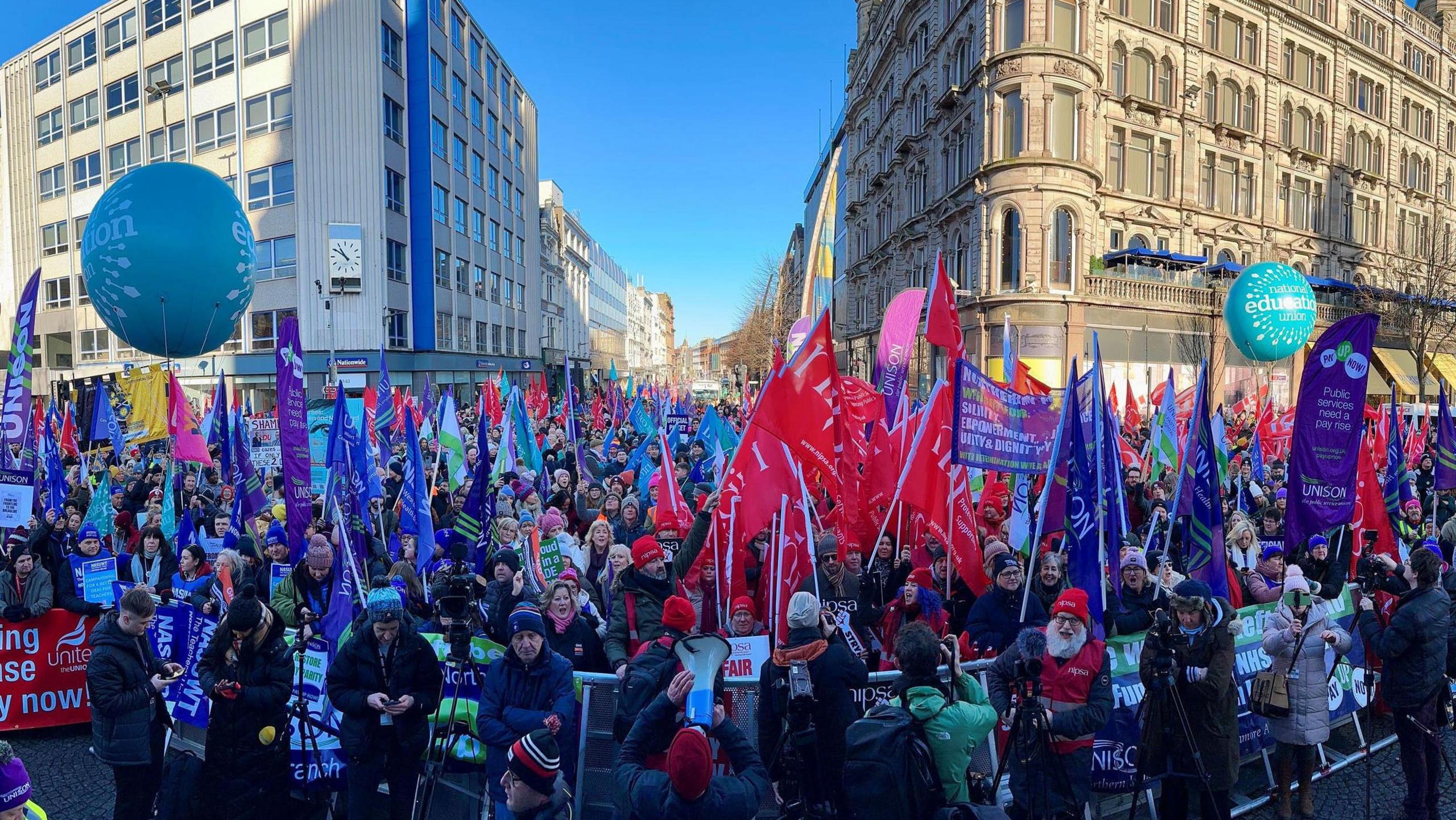 A wide shot of a large crowd of health workers on strike - they are holding up various banners. Most of the flags are either purple and green or red and white.