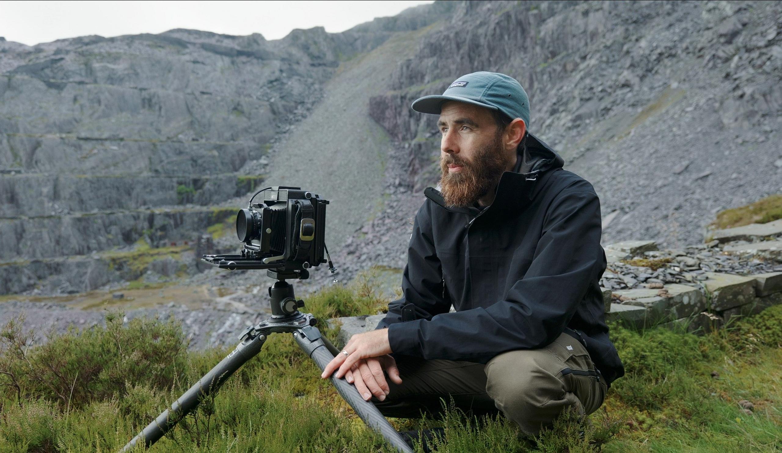 Kyle McDougall is wearing a blue baseball hat, navy raincoat, khaki trousers and has a bushy beard. Hi is crouched down on grass in a slate quarry with slate rising up behind him. 