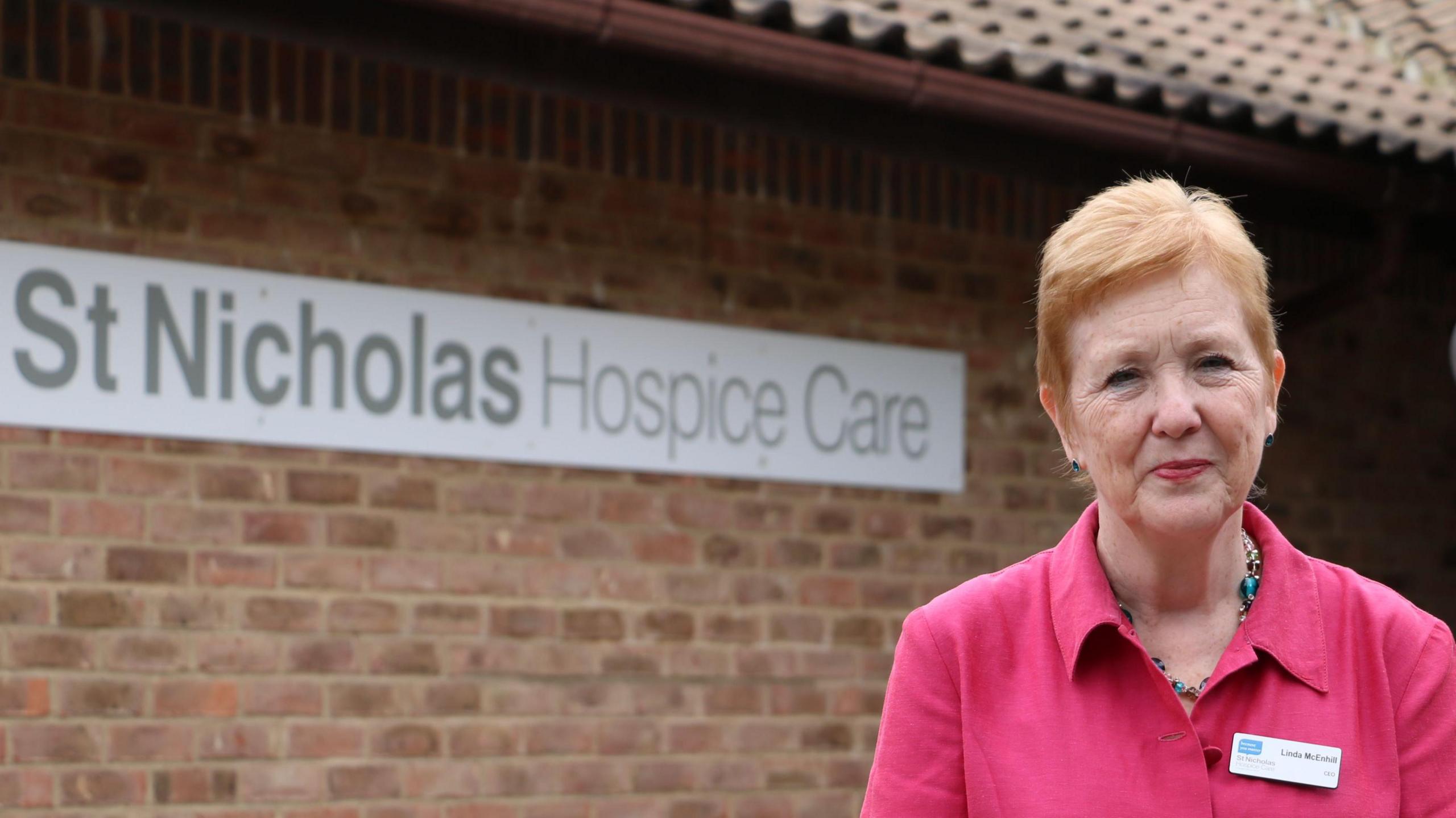 A photo of Linda McEnhill. She is facing and smiling at the camera. She is standing outside St Nicholas Hospice in Bury St Edmunds. She is wearing a bright pink shirt with a name tag as well as a necklace around her neck. She has short ginger hair